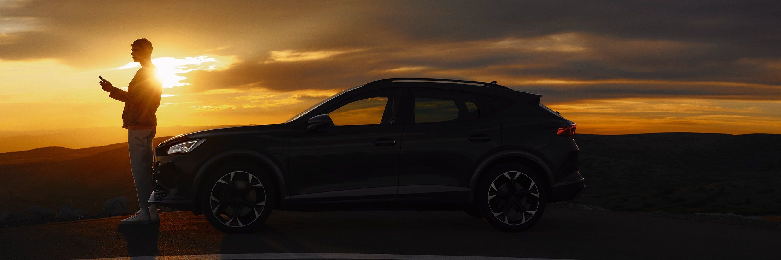 Osoba stojí vedľa zaparkovaného automobilu na kraji cesty pri západe slnka. Obloha je pokrytá tmavými mrakmi, ktoré vytvárajú dramatický efekt svetla. 