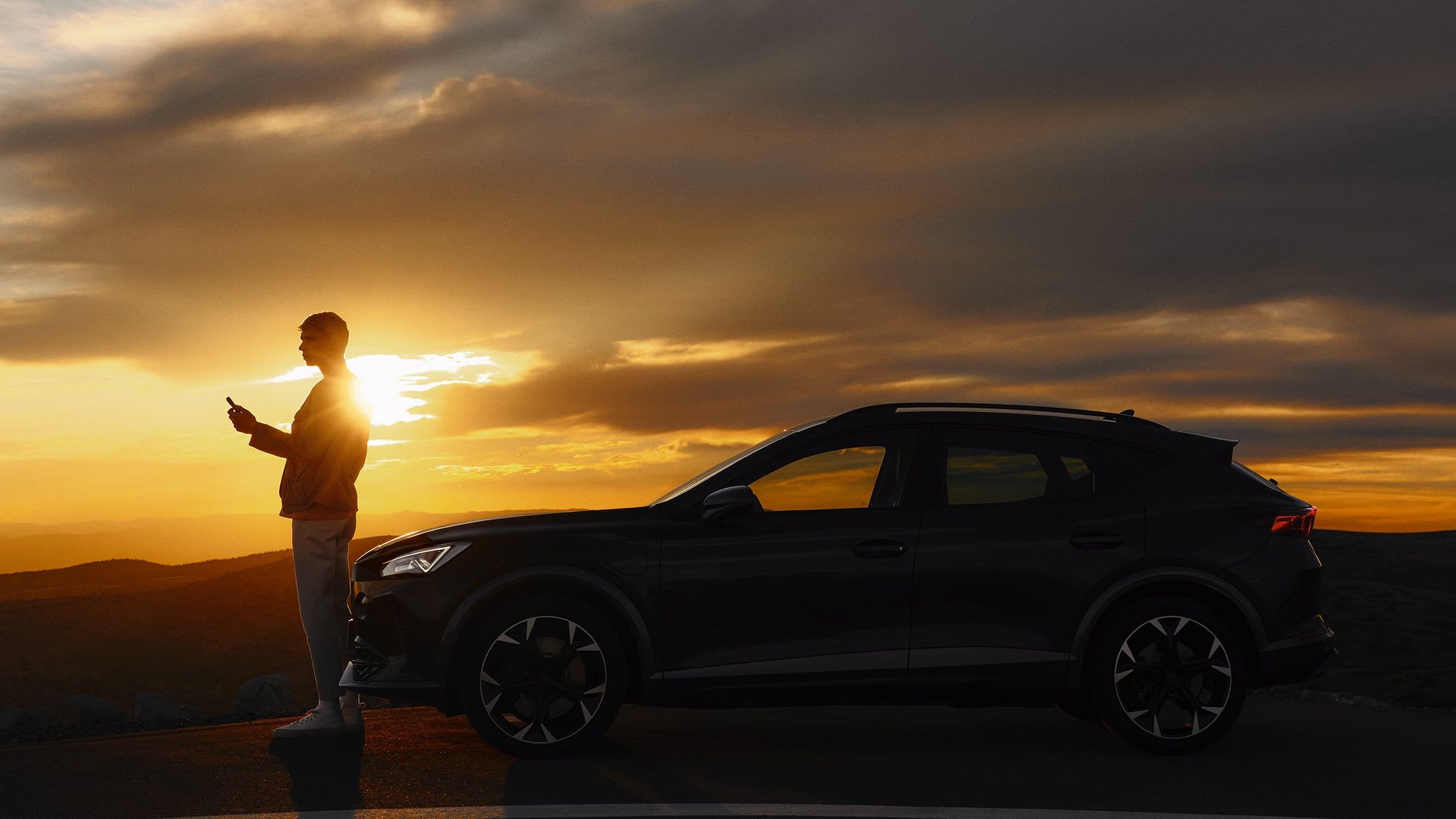 Osoba stojí vedľa zaparkovaného automobilu na kraji cesty pri západe slnka. Obloha je pokrytá tmavými mrakmi, ktoré vytvárajú dramatický efekt svetla. 