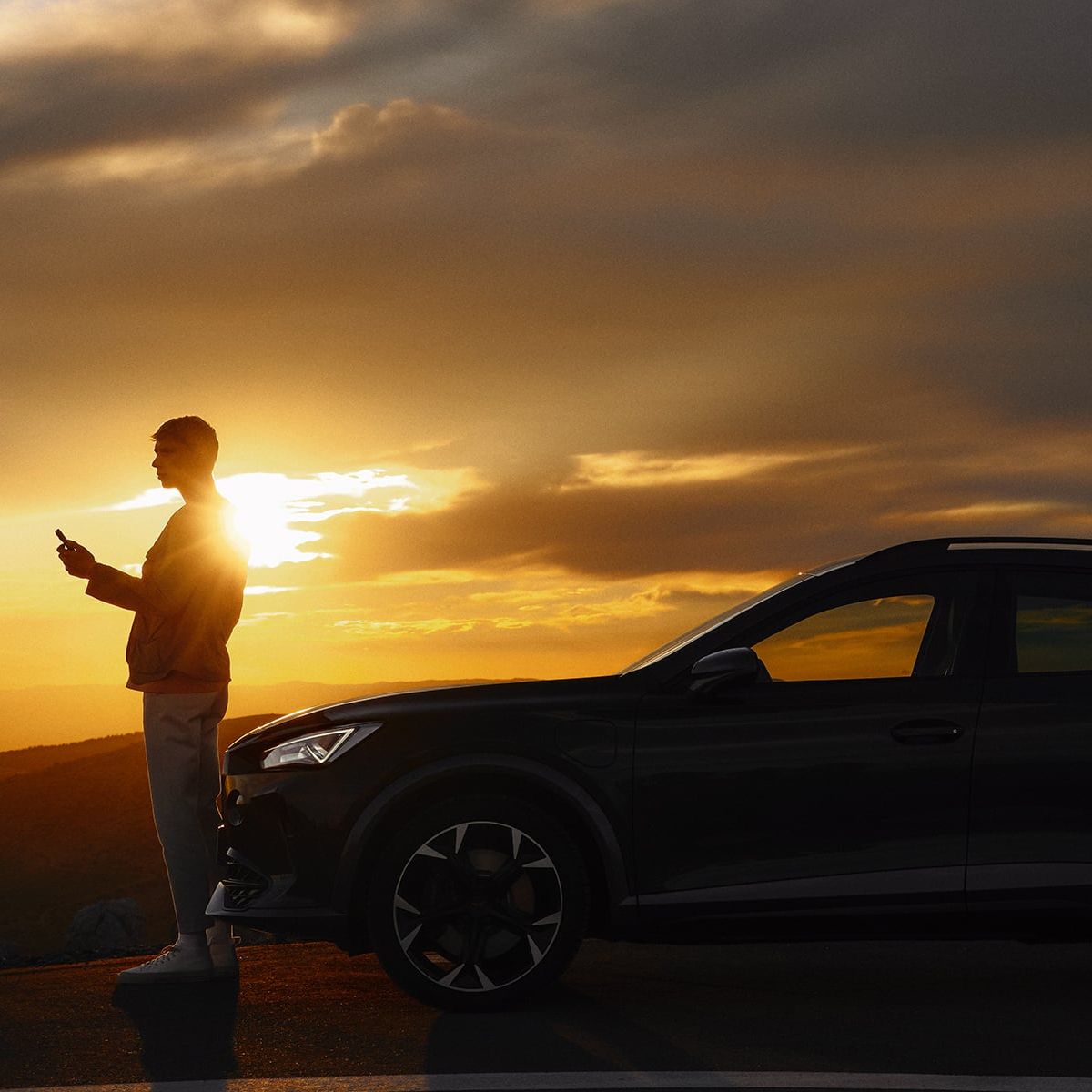 Osoba stojí vedľa zaparkovaného automobilu na kraji cesty pri západe slnka. Obloha je pokrytá tmavými mrakmi, ktoré vytvárajú dramatický efekt svetla. 