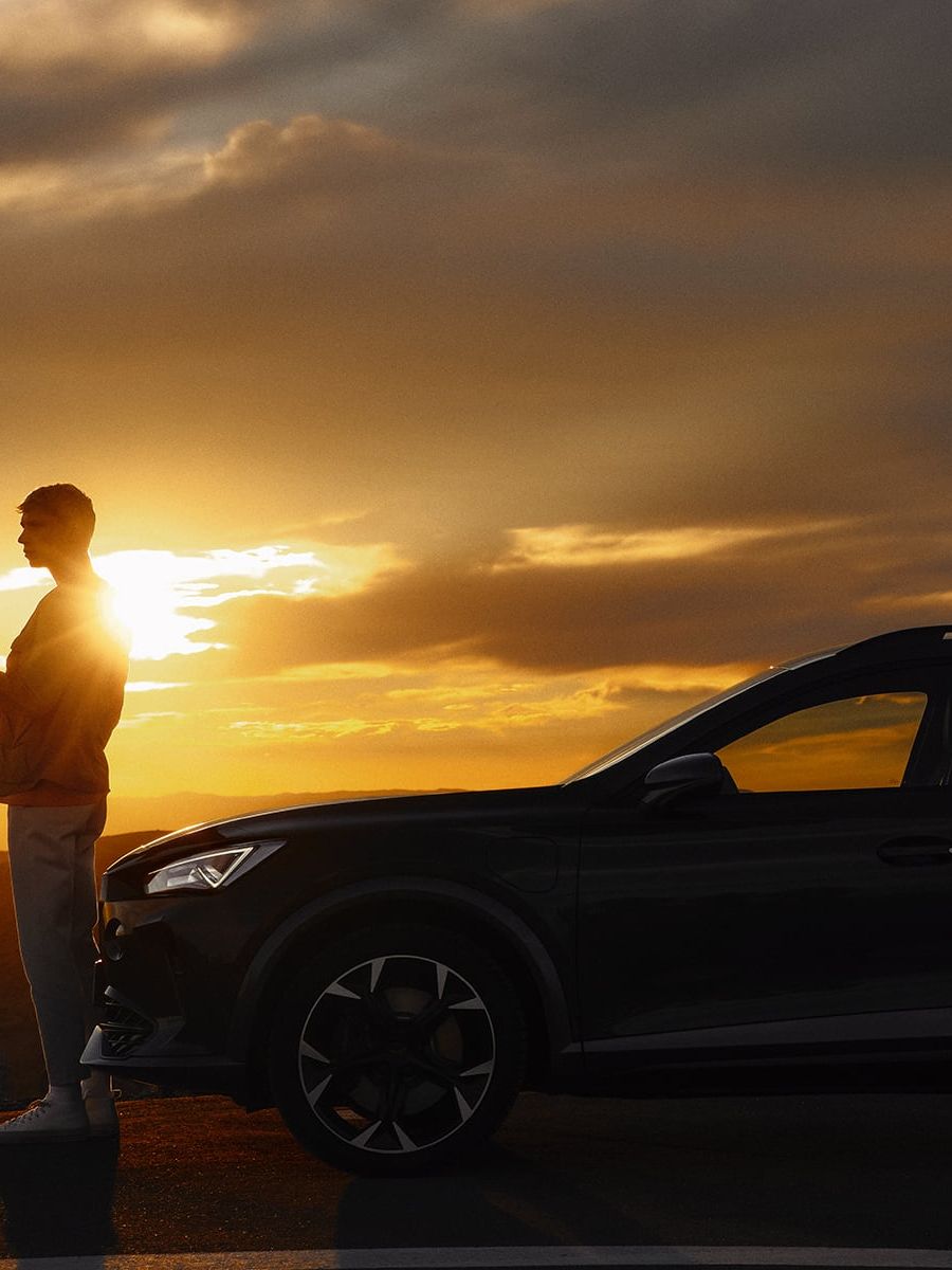 Osoba stojí vedľa zaparkovaného automobilu na kraji cesty pri západe slnka. Obloha je pokrytá tmavými mrakmi, ktoré vytvárajú dramatický efekt svetla. 