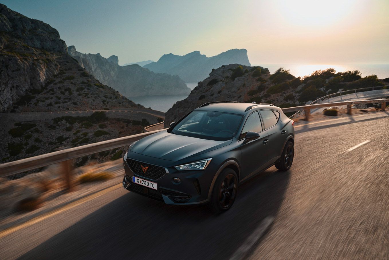 Športové SUV jazdí po horskej ceste za slnečného dňa. Okolo cesty sú vysoké skaly a v pozadí je výhľad na more. Atmosféra vyjadruje rýchlosť a slobodu.