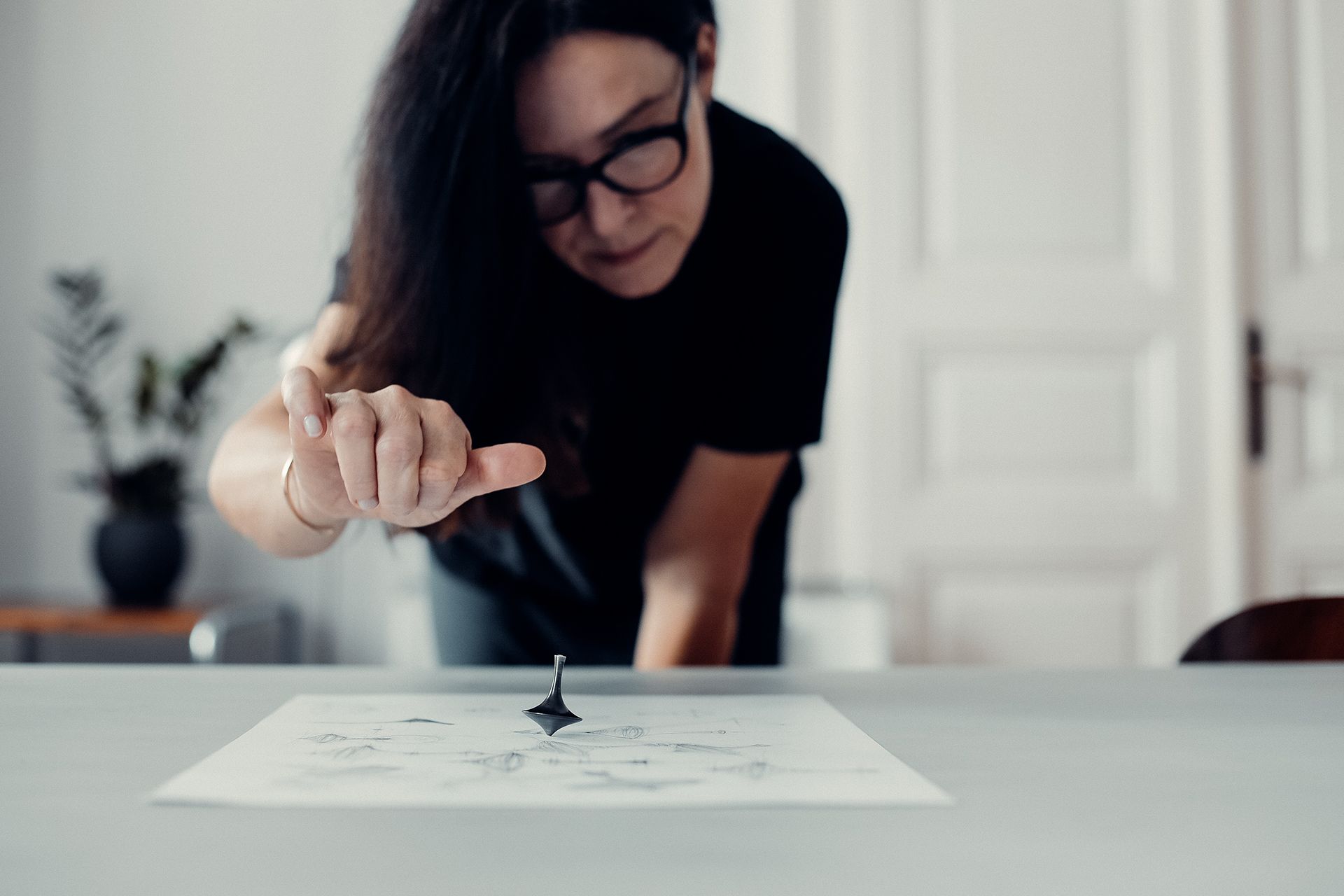 Žena ukazuje na točiaci sa kovový predmet položený na papieri. V pozadí je miestnosť s bielymi dverami a rastlinami.