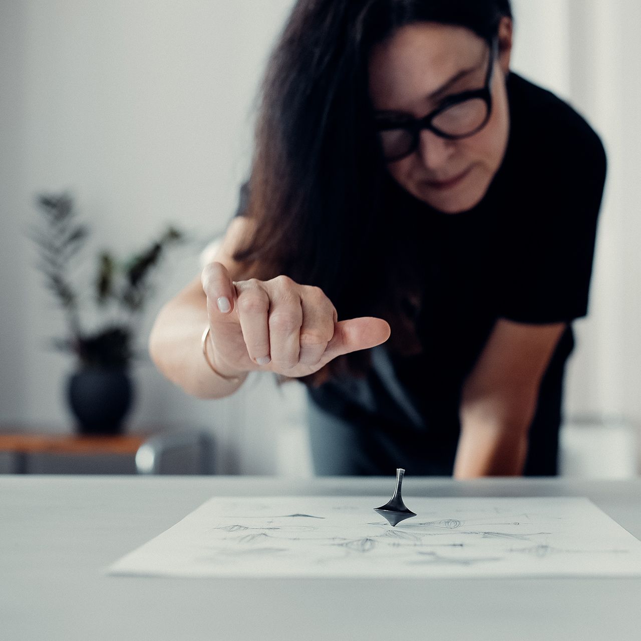 Žena ukazuje na točiaci sa kovový predmet položený na papieri. V pozadí je miestnosť s bielymi dverami a rastlinami.