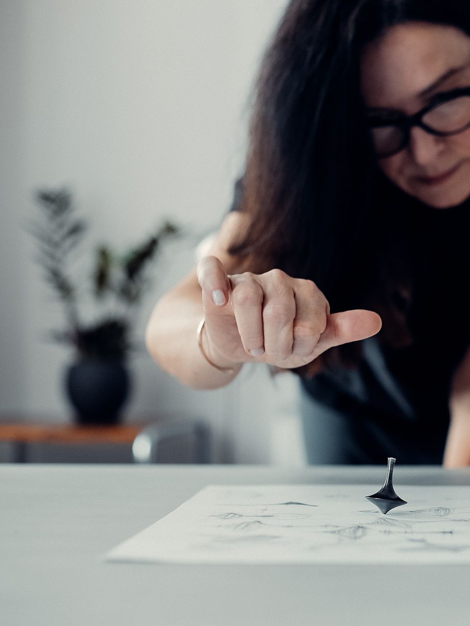 Žena ukazuje na točiaci sa kovový predmet položený na papieri. V pozadí je miestnosť s bielymi dverami a rastlinami.