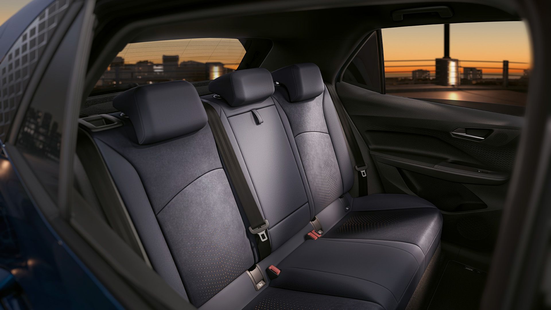 Interiér zadných sedadiel moderného automobilu. Sedadlá sú čalúnené v tmavej látke a majú bezpečnostné pásy. V pozadí je vidieť mesto pri západe slnka.