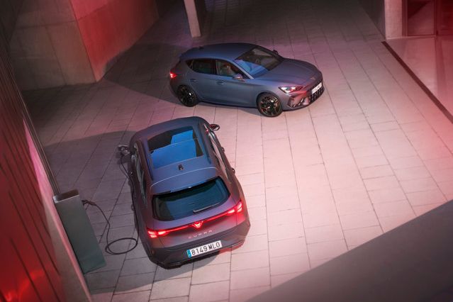 Nočný výhľad zhora na dve zaparkované autá na osvetlenom betónovom nádvorí. Jedno z áut je pripojené k nabíjacej stanici. Okolie je obklopené modernými budovami so svetelnými efektami.