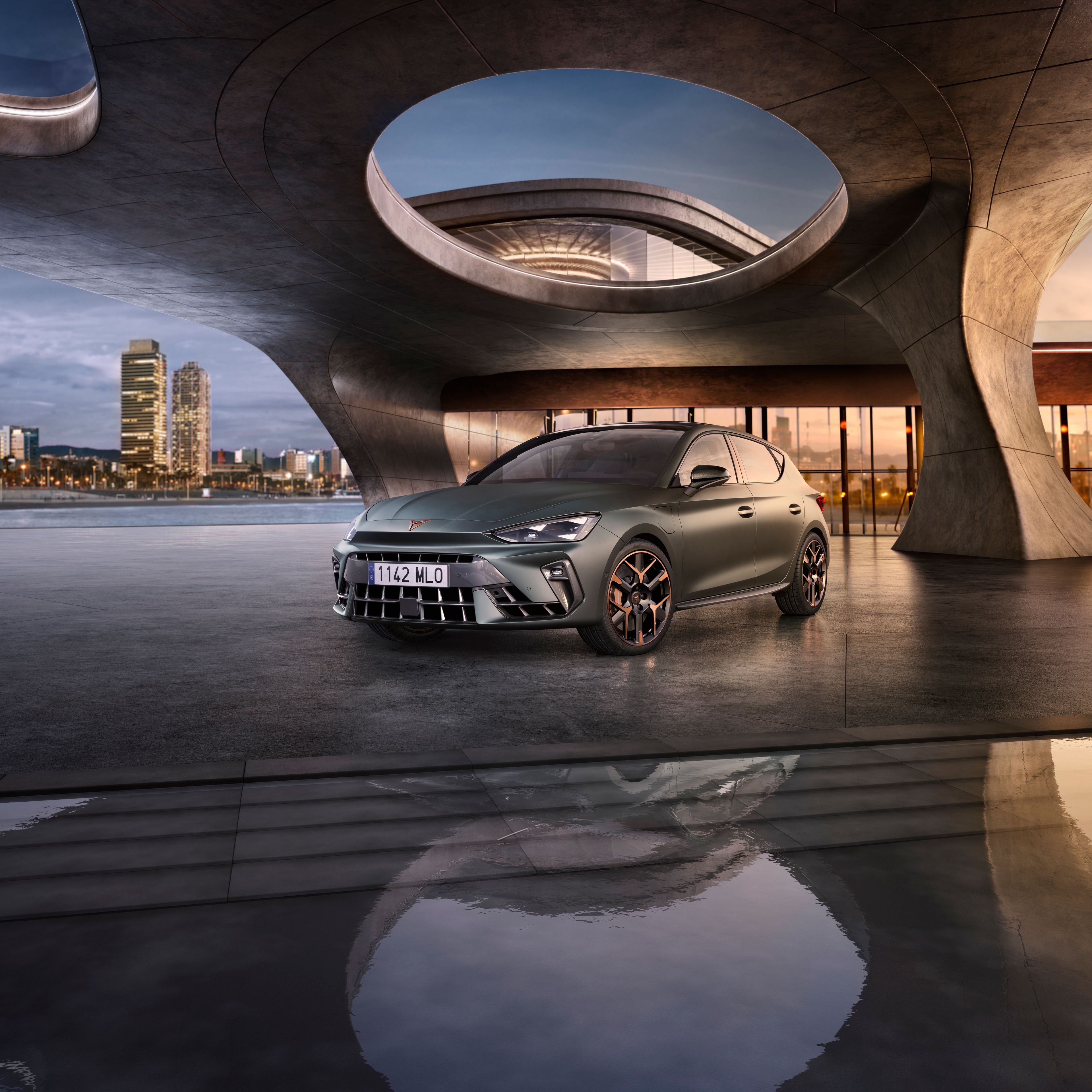 CUPRA Leon stojí pri modernej stavbe pod strechou s kruhovými sklenenými otvormi, ktorá stojí pri mori a v pozadí vidieť mesto.