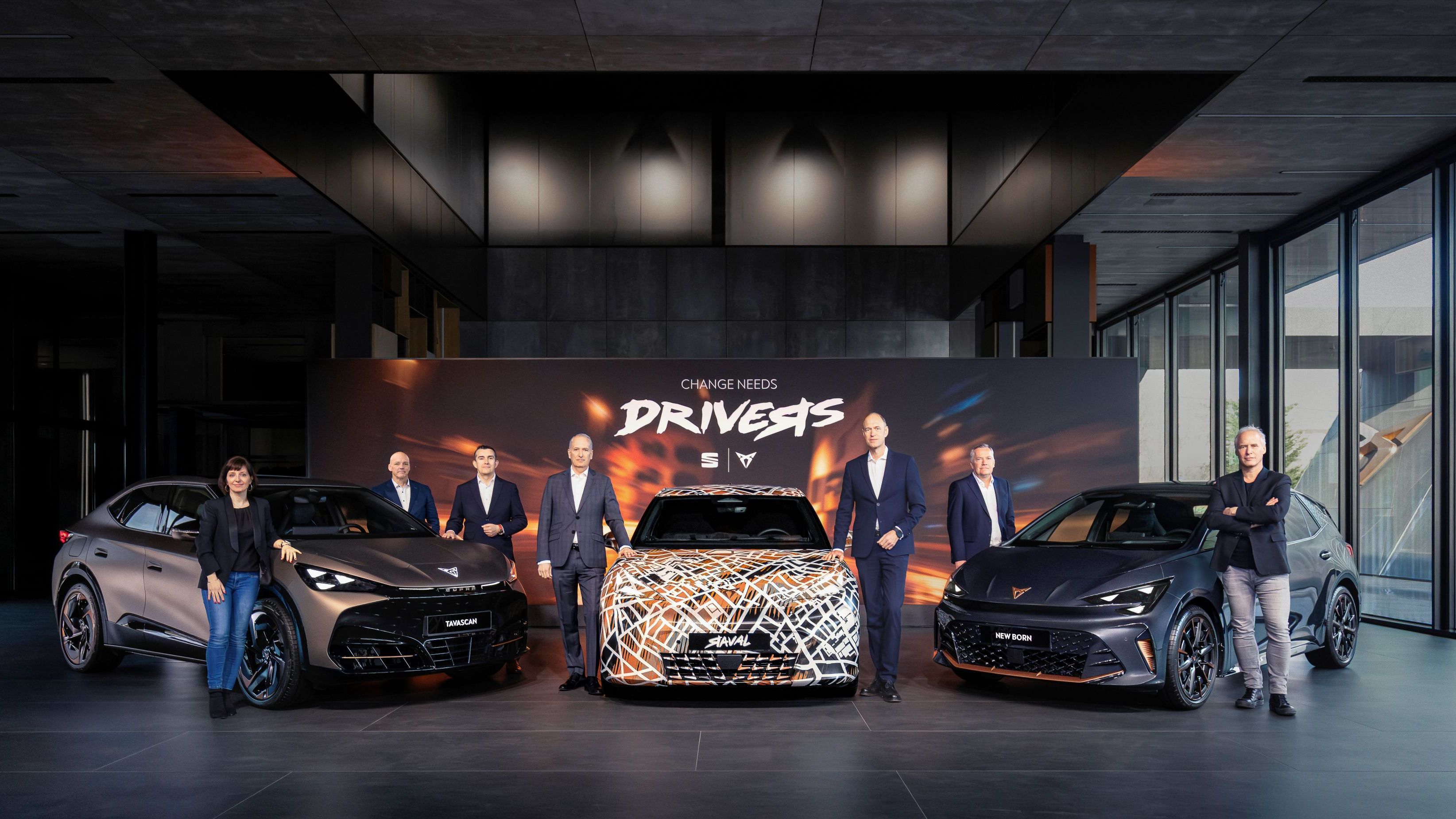Tri modeli CUPRA zaparkované v modernom, dizajnovom showroome. Vedľa vozidiel stoja manažéri. Za vozidlami je veľký reklamný panel s nápisom "Change needs drivers." a logami SEAT a CUPRA. 
