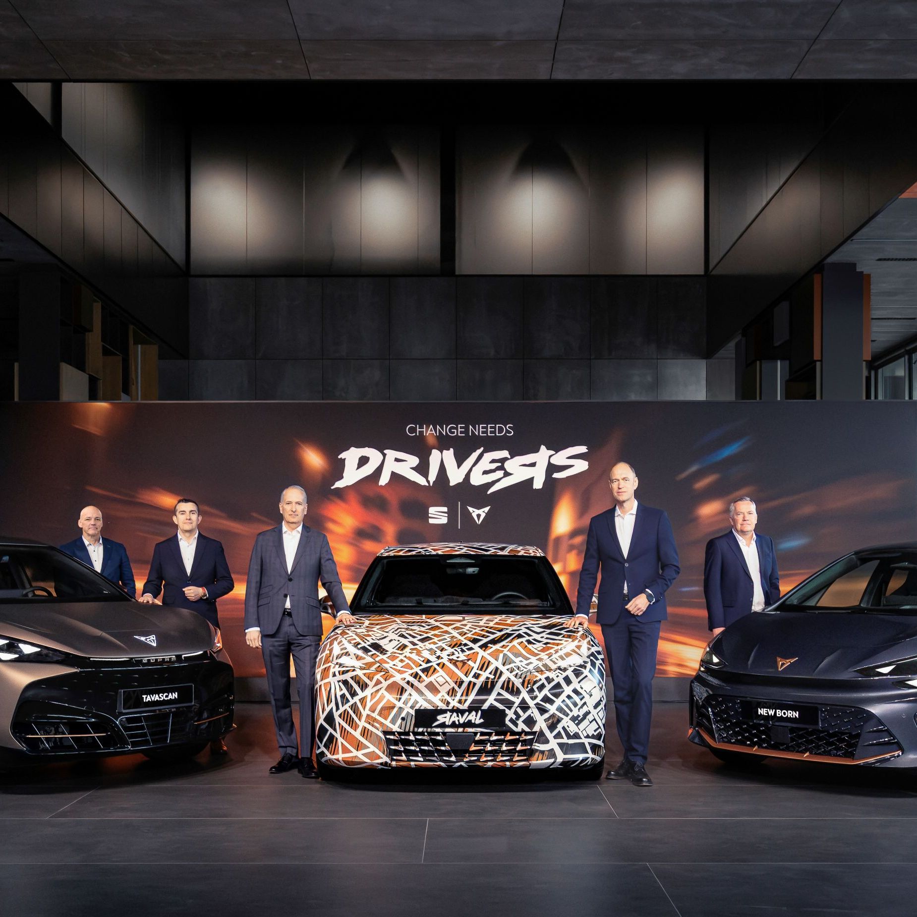 Tri modeli CUPRA zaparkované v modernom, dizajnovom showroome. Vedľa vozidiel stoja manažéri. Za vozidlami je veľký reklamný panel s nápisom "Change needs drivers." a logami SEAT a CUPRA. 