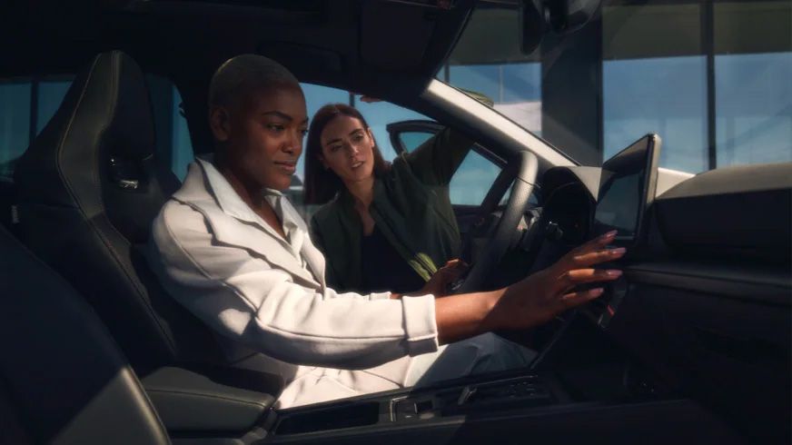 Dve osoby v a pri luxusnom vozidle. Jedna žena sedí za volantom a pozerá sa na displej infotainment, zatiaľ čo druhá sa tiež pozerá na displeji. Interiér vozidla je moderný a elegantný.