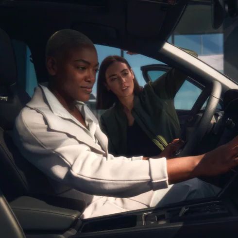 Dve osoby v a pri luxusnom vozidle. Jedna žena sedí za volantom a pozerá sa na displej infotainment, zatiaľ čo druhá sa tiež pozerá na displeji. Interiér vozidla je moderný a elegantný.