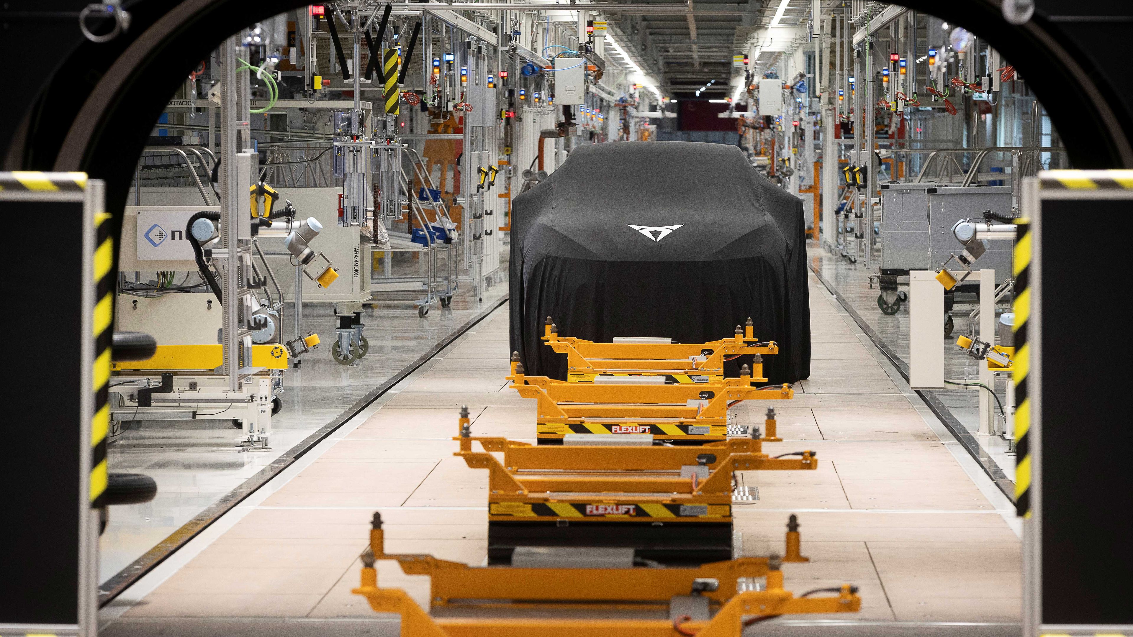 Automobil zahalený čiernou plachtou stojí vo výrobnej hale plnej priemyselných zariadení. Pred vozidlom sú žlté palety na podlahových koľajniciach. V stene je veľký otvor, cez ktorý je vidieť časť výrobnej linky.