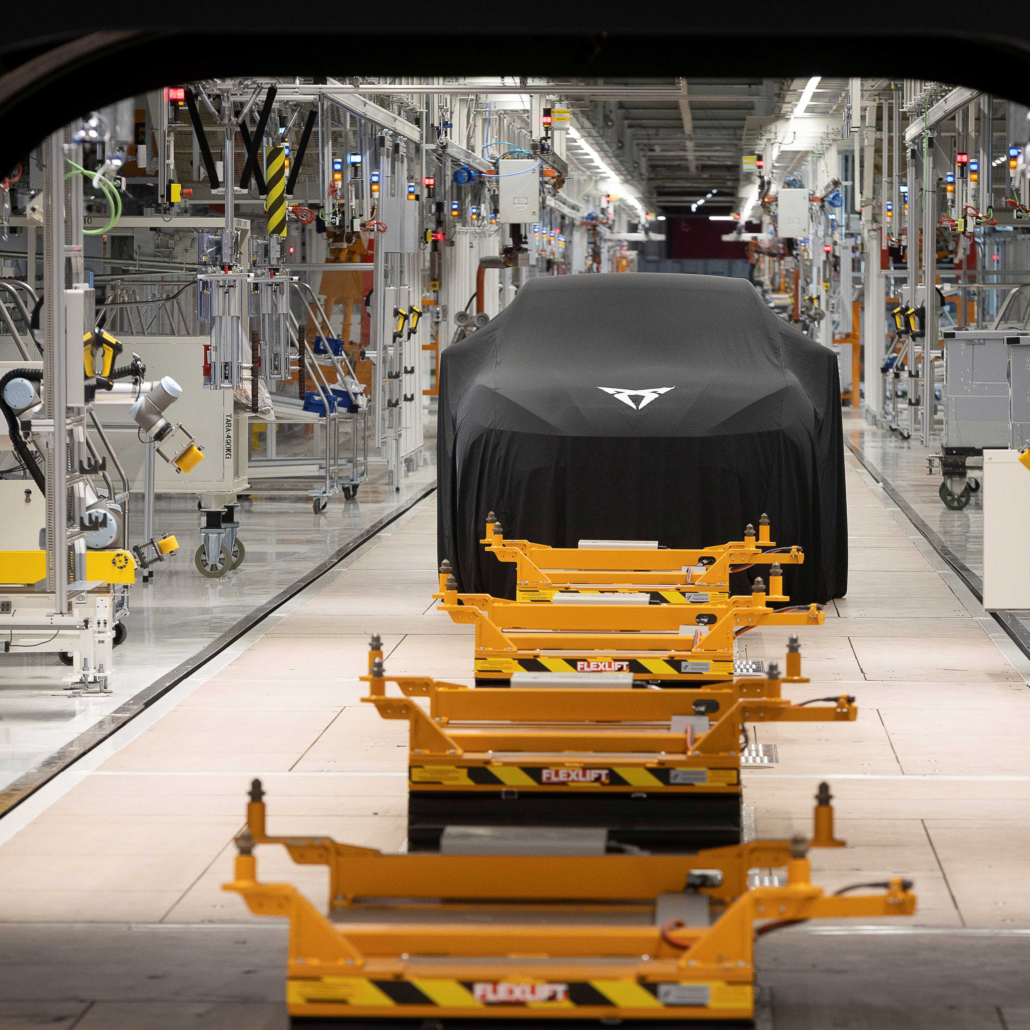 Automobil zahalený čiernou plachtou stojí vo výrobnej hale plnej priemyselných zariadení. Pred vozidlom sú žlté palety na podlahových koľajniciach. V stene je veľký otvor, cez ktorý je vidieť časť výrobnej linky.