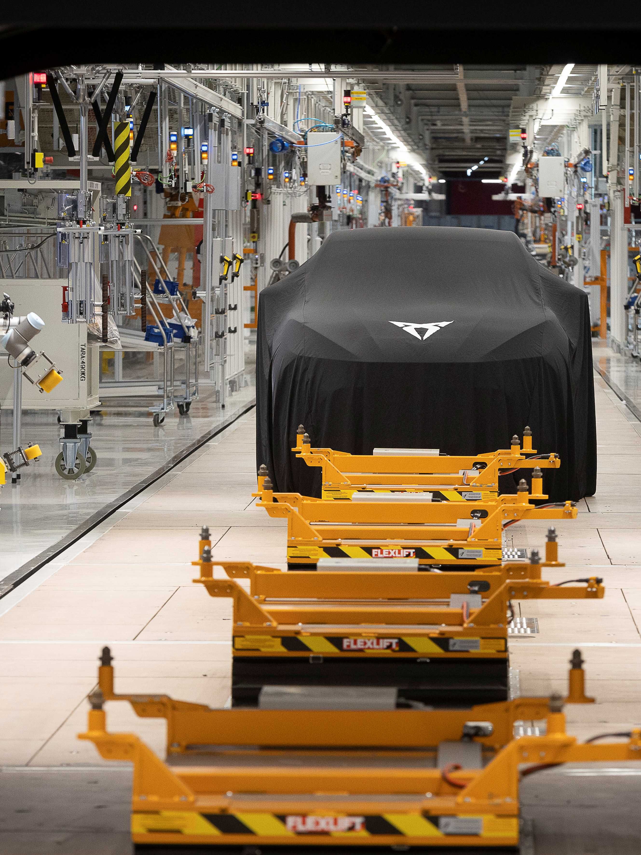 Automobil zahalený čiernou plachtou stojí vo výrobnej hale plnej priemyselných zariadení. Pred vozidlom sú žlté palety na podlahových koľajniciach. V stene je veľký otvor, cez ktorý je vidieť časť výrobnej linky.