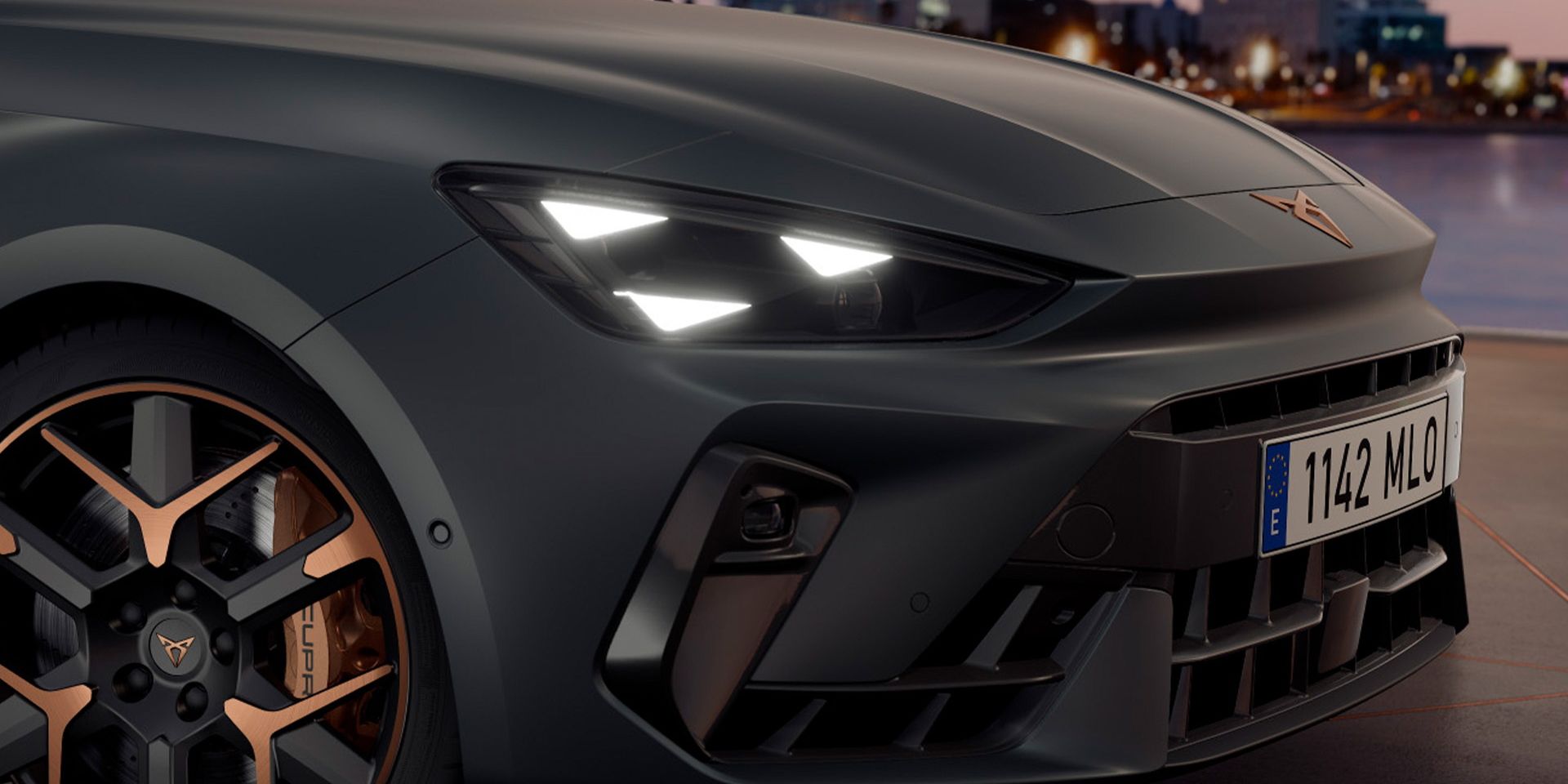 Detailný záber na prednú časť športového vozidla so zapnutými svetlometmi. V pozadí je vidieť mesto pri vode počas západu slnka. Auto má štýlové kolesá s medenými akcentmi.