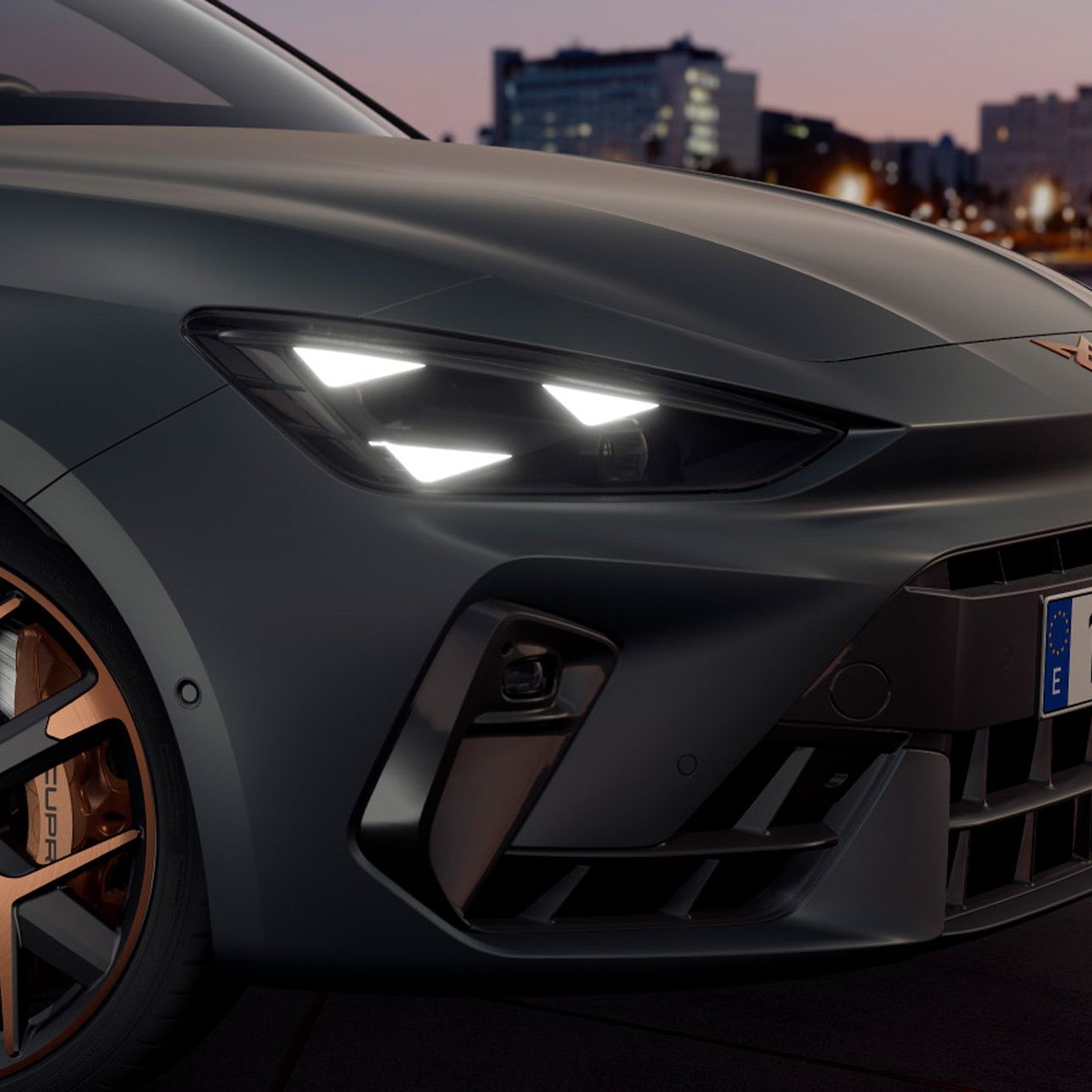 Detailný záber na prednú časť športového vozidla so zapnutými svetlometmi. V pozadí je vidieť mesto pri vode počas západu slnka. Auto má štýlové kolesá s medenými akcentmi.