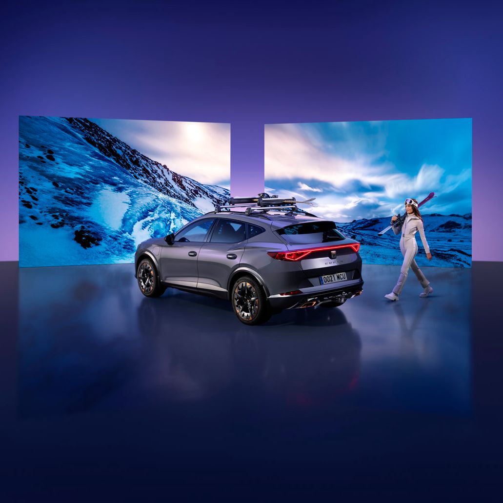 Šedé SUV s lyžami na strešnom nosiči stojí pred veľkými obrazovkami zobrazujúcimi zasneženú horskú krajinu. Vedľa auta kráča žena oblečená v lyžiarskom oblečení s lyžami na ramene. Pozadie je tmavofialové.