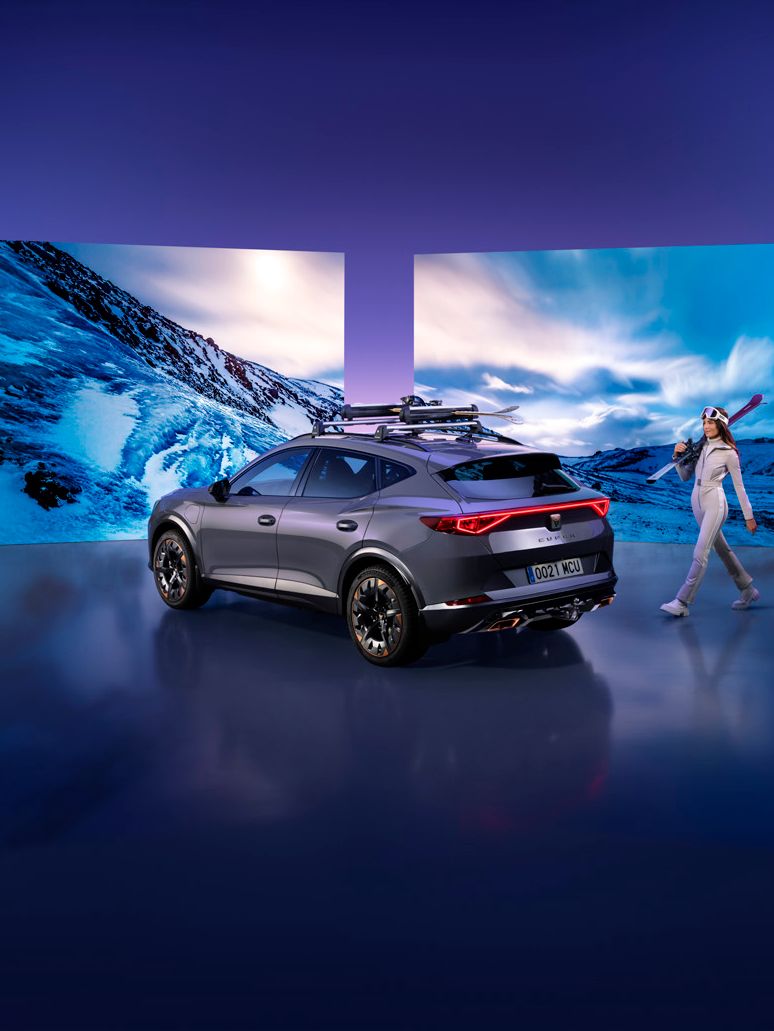 Šedé SUV s lyžami na strešnom nosiči stojí pred veľkými obrazovkami zobrazujúcimi zasneženú horskú krajinu. Vedľa auta kráča žena oblečená v lyžiarskom oblečení s lyžami na ramene. Pozadie je tmavofialové.