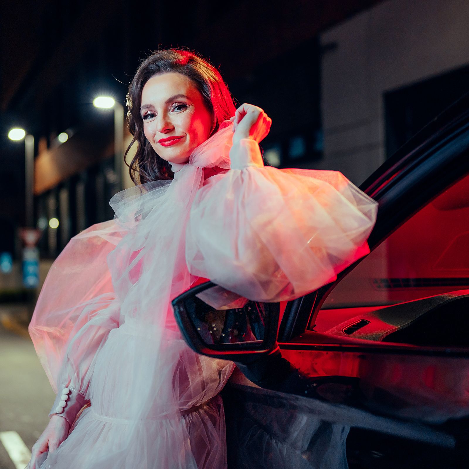 Žena stojí opretá o auto na ulici v noci, zahalená v priehľadnom bielom oblečení. Štýl fotografie je dramatický, zvýraznený červeným osvetlením od auta.