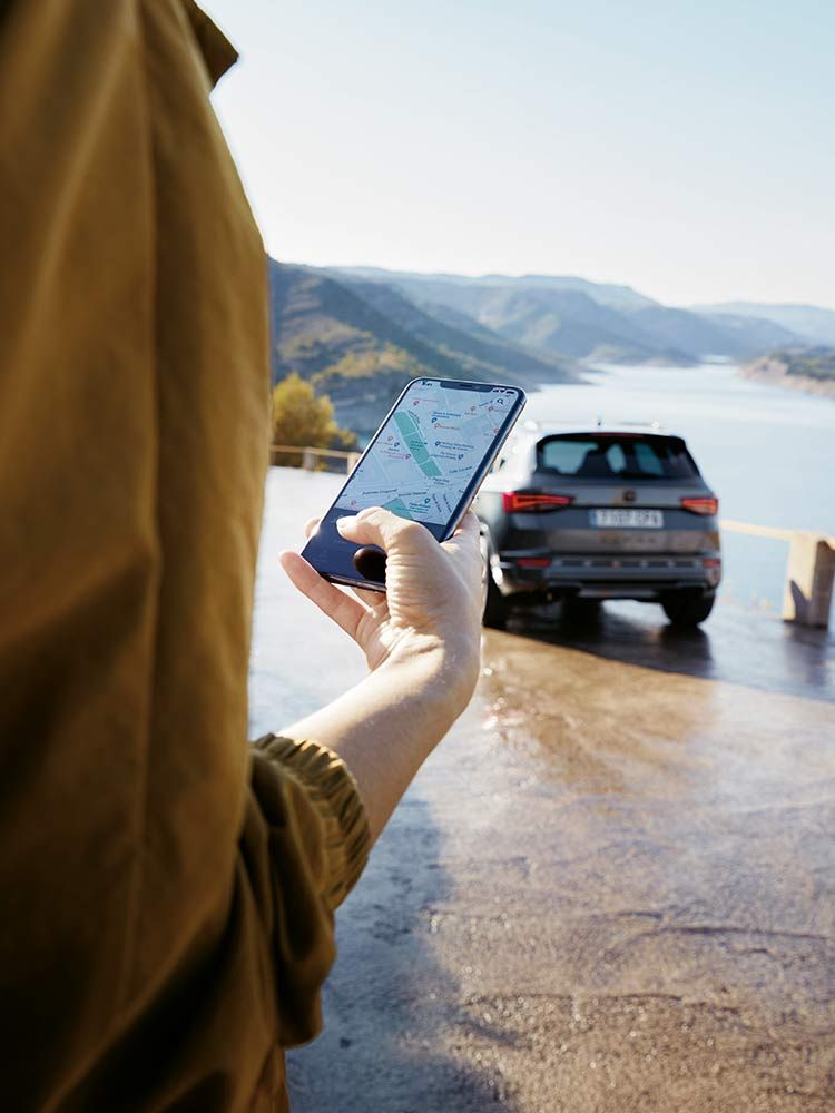 Osoba držiaca mobilný telefón s otvorenou navigačnou aplikáciou. V pozadí zaparkované auto pri jazere s kopcami. Scéna je zrejme pri ceste s nádherným výhľadom na prírodu.