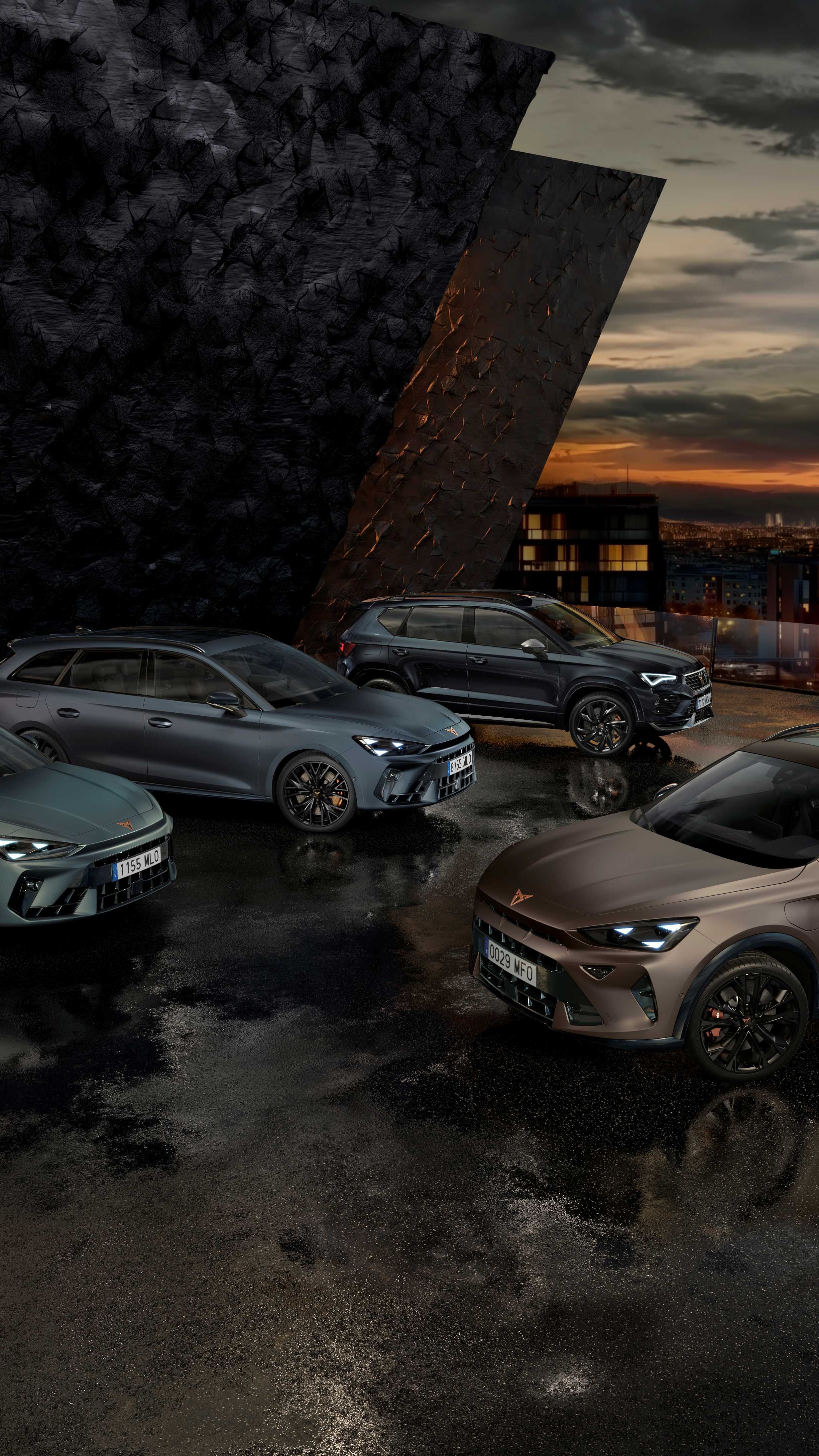 Na obrázku sú štyri autá značky Cupra zaparkované vedľa moderného architektonického objektu počas súmraku. Každé auto má športový dizajn a tmavé metalické farby. Pozadie tvorí mestská scenéria s budovami a večerným osvetlením.