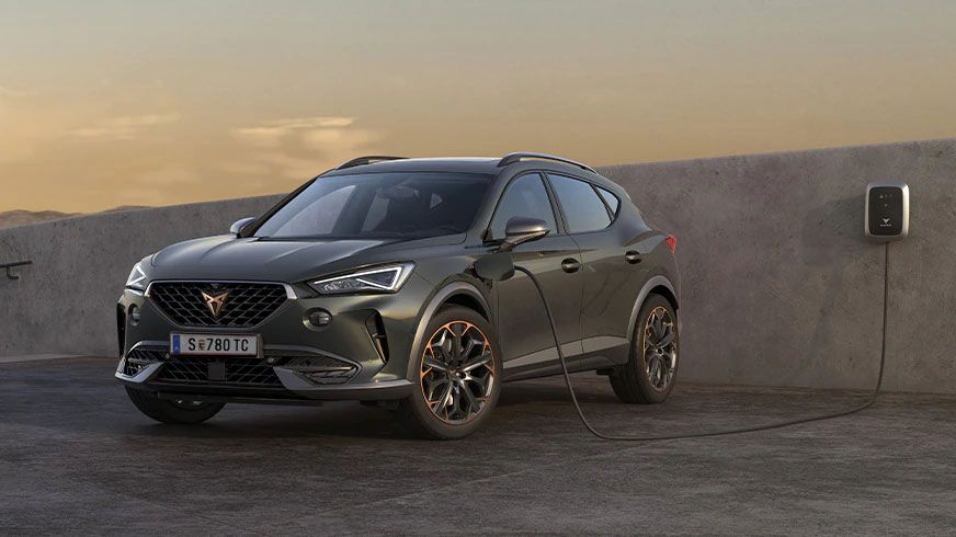 Moderné sivé SUV je pripojené na nabíjaciu stanicu pri betónovej stene. Auto má elegantný dizajn s výraznými svetlami a veľkými kolesami. Nabíjacia stanica je umiestnená na stene a zapojená do vozidla.