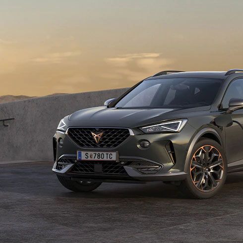 Moderné sivé SUV je pripojené na nabíjaciu stanicu pri betónovej stene. Auto má elegantný dizajn s výraznými svetlami a veľkými kolesami. Nabíjacia stanica je umiestnená na stene a zapojená do vozidla.