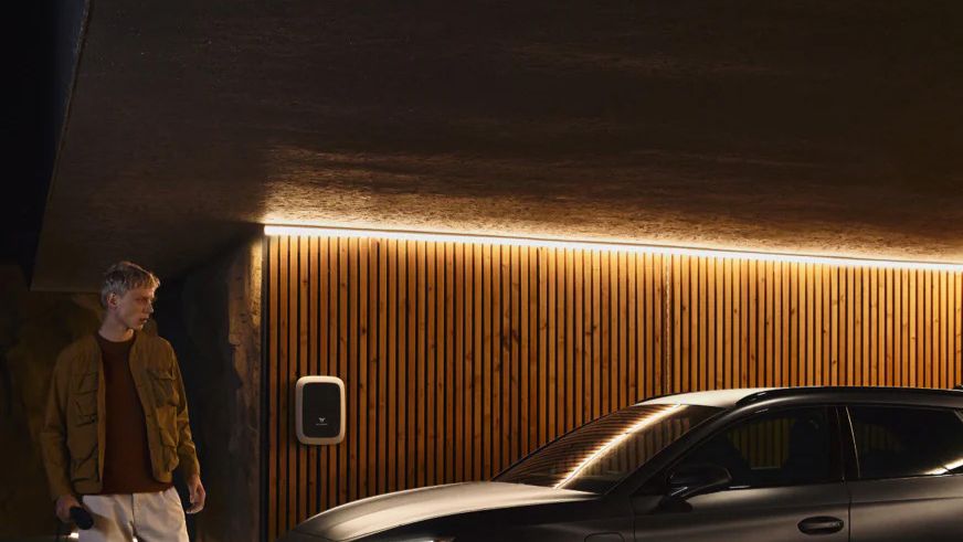 Osoba stojí pri zaparkovanom striebornom vozidle, ktoré sa nabíja na nabíjacej stanici. Nabíjacia stanica je integrovaná na drevenú stenu domu, ktorá je osvetlená bielym svetlom zhora.