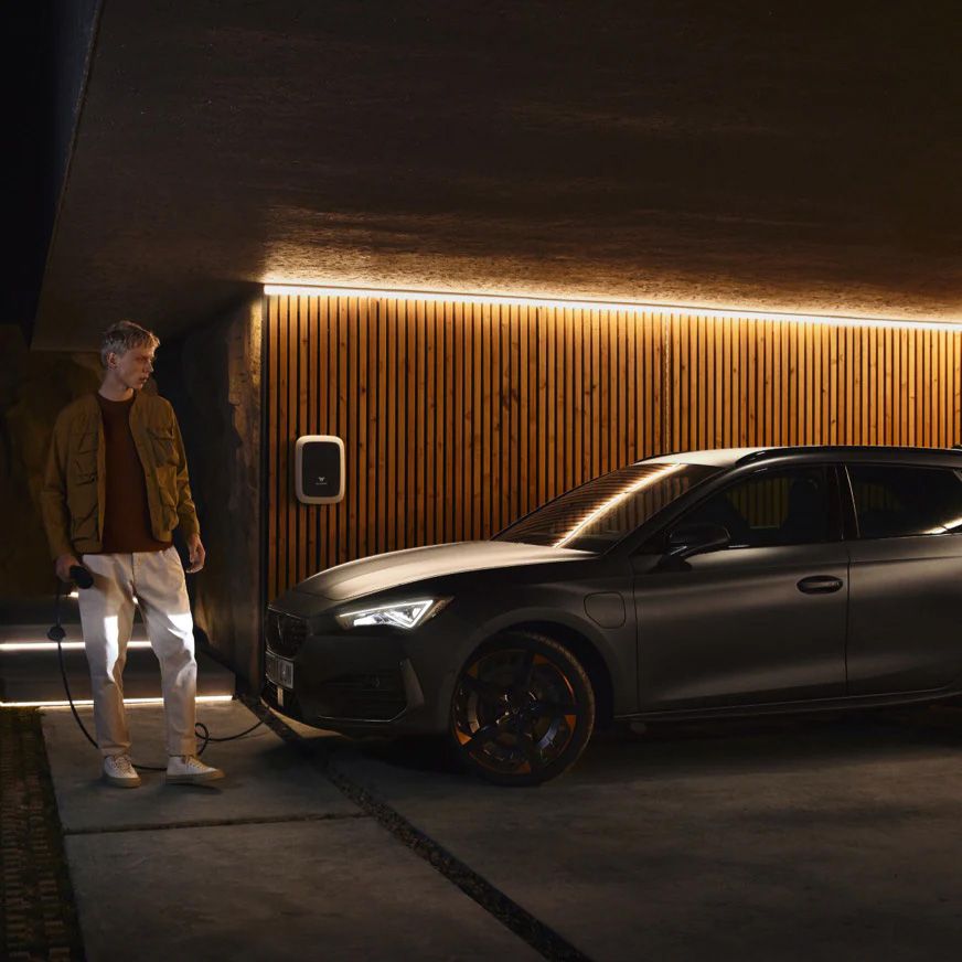Osoba stojí pri zaparkovanom striebornom vozidle, ktoré sa nabíja na nabíjacej stanici. Nabíjacia stanica je integrovaná na drevenú stenu domu, ktorá je osvetlená bielym svetlom zhora.