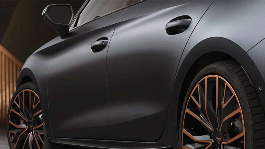 Detailný záber na bočné čierne kolesá športového auta s bronzovými prvkami. Automobil má elegantný tmavý matný povrch. Na pozadí je drevené zábradlie.