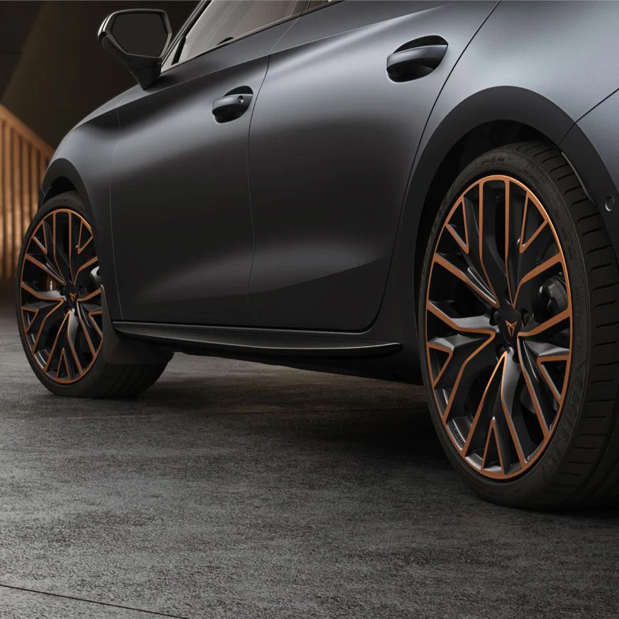 Detailný záber na bočné čierne kolesá športového auta s bronzovými prvkami. Automobil má elegantný tmavý matný povrch. Na pozadí je drevené zábradlie.