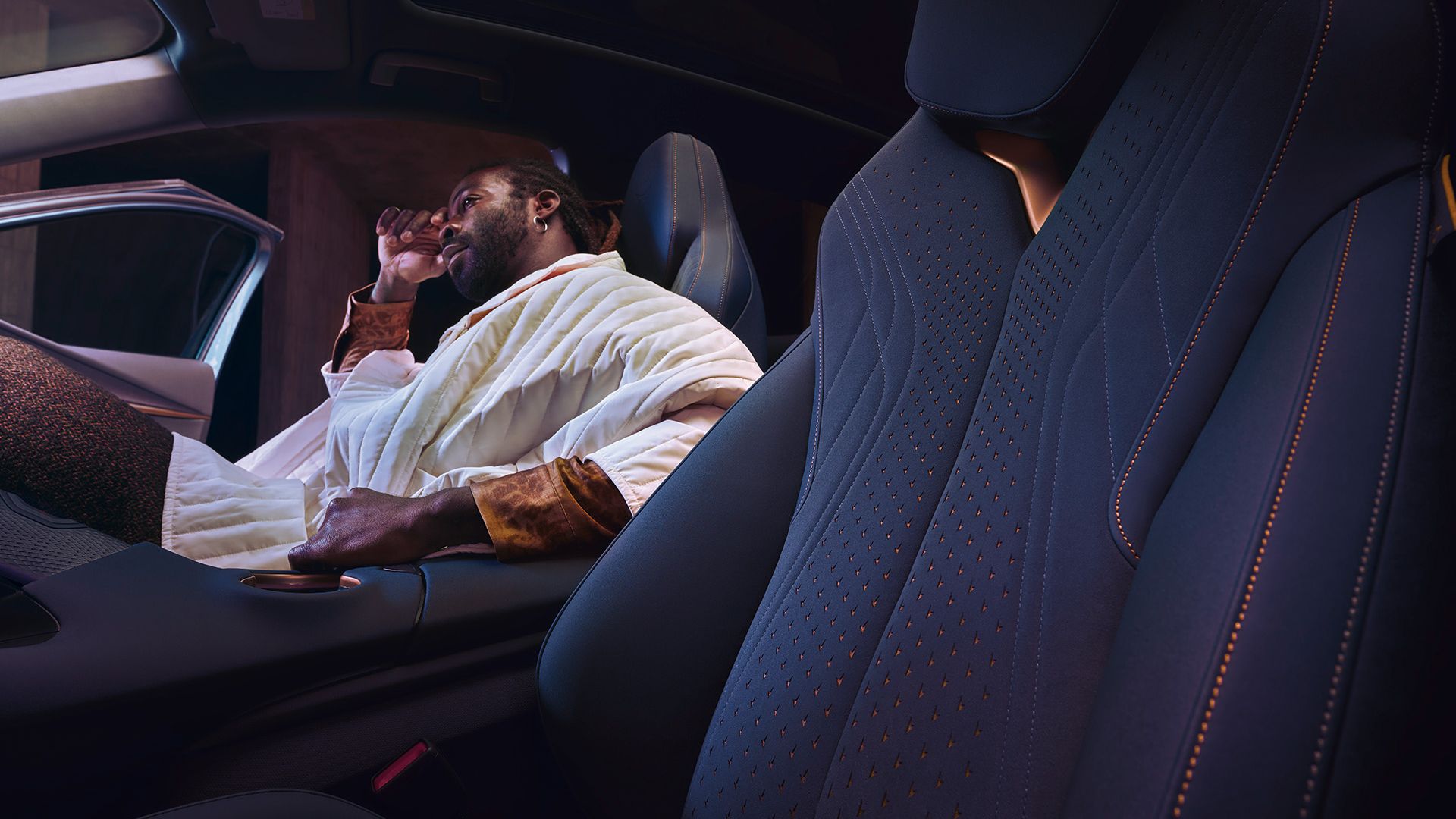 Muž sedí na sedadle spolujazdca a má na sebe biely prešívaný kabát. Vozidlo má moderný interiér s čiernymi sedadlami s červeným prešívaním.