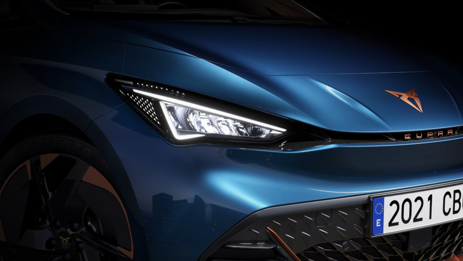 Detailný záber predných partií tmavomodrého športového auta s výraznými LED svetlometmi a španielskou poznávacou značkou. Na kapote je logo značky Cupra. 