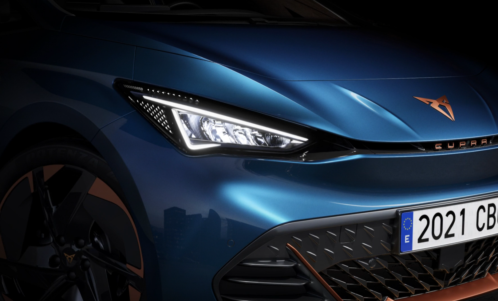 Detailný záber predných partií tmavomodrého športového auta s výraznými LED svetlometmi a španielskou poznávacou značkou. Na kapote je logo značky Cupra. 