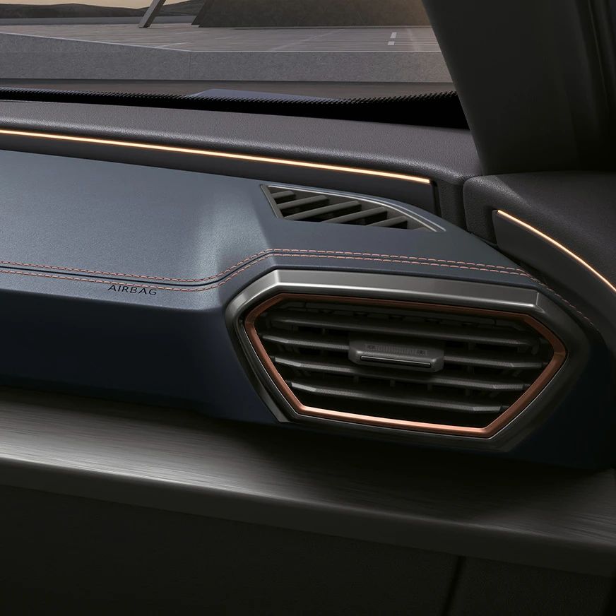 Detail na interiér auta, konkrétne na pravú stranu palubnej dosky s ventilačným otvorom. Na palubnej doske je vidieť nápis "Airbag". Pozadie je tmavé a palubná doska má elegantné prešívanie.