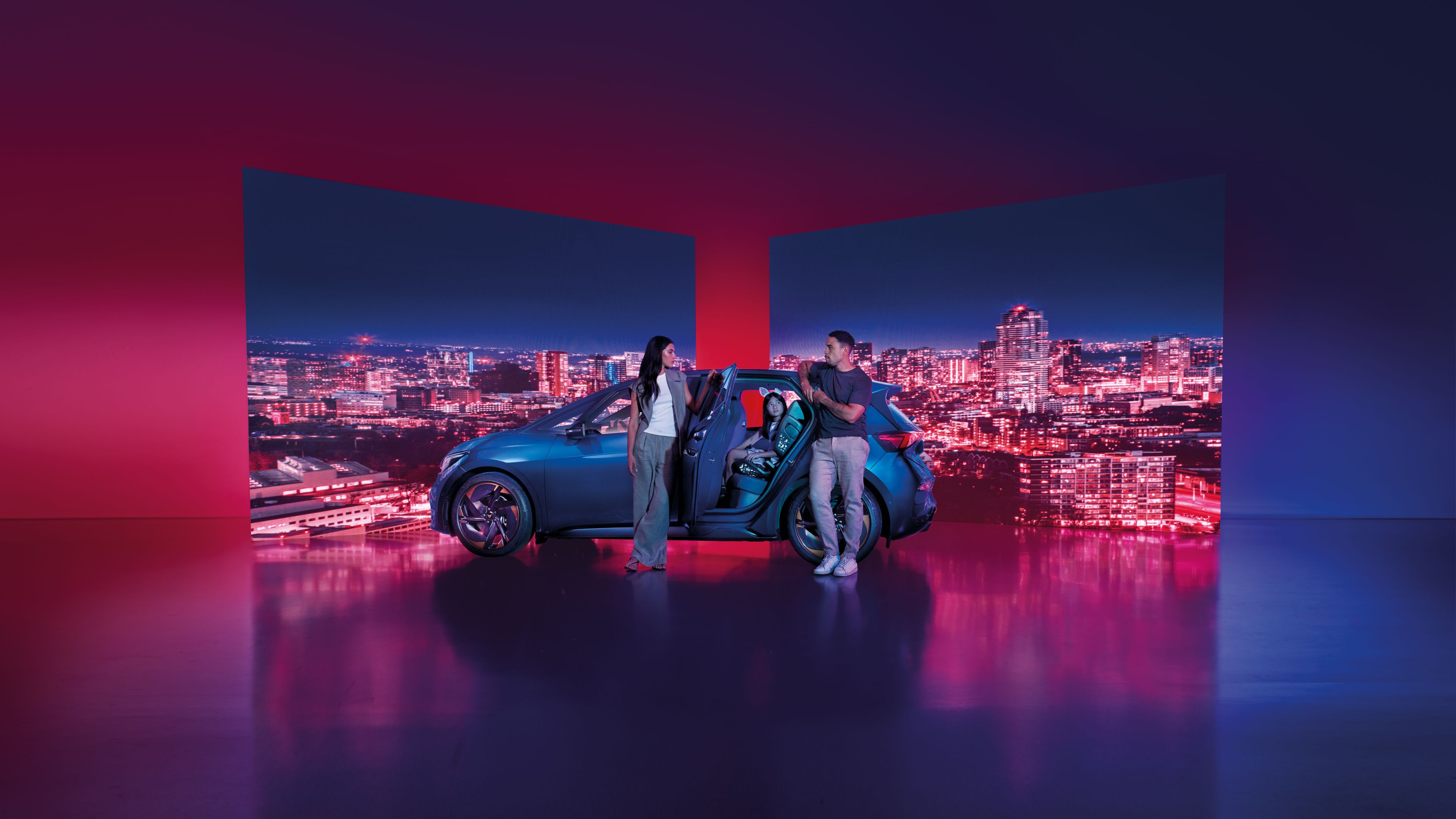 Dvaja ľudia stoja vedľa modrého auta v miestnosti s osvetleným pozadím, ktoré tvoria dve obrazovky s nočným mestom s osvetlenými budovami.