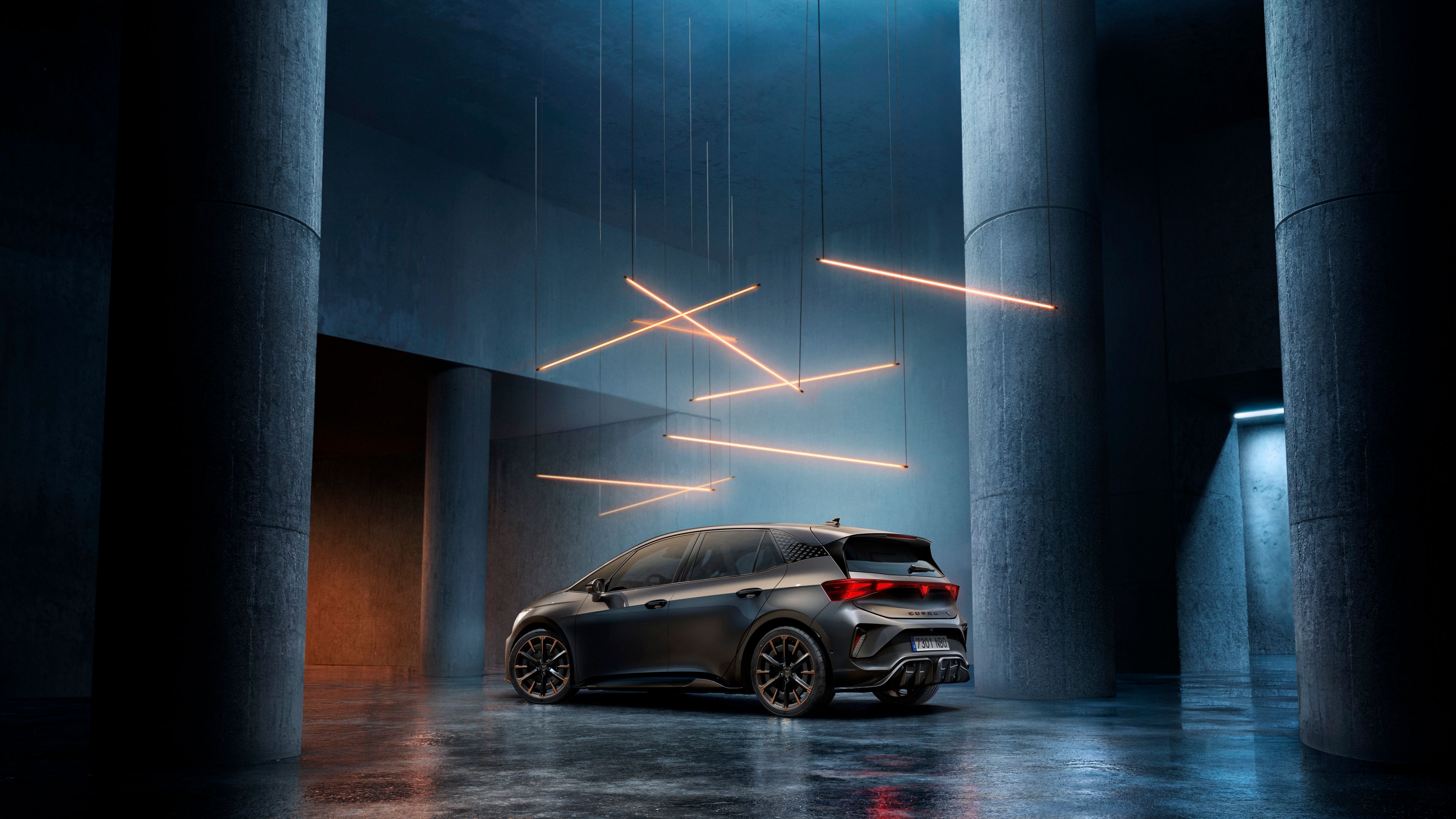 CUPRA Born stojí v štýlovo osvetlenom priemyselnom priestore s betónovými stenami a stĺpmi. Zo stropu visí niekoľko lineárnych svetelných zdrojov, ktoré vytvárajú elegantnú atmosféru. Vozidlo je vidieť zo zadnej strany a odráža svetlo na lesklej podlahe.