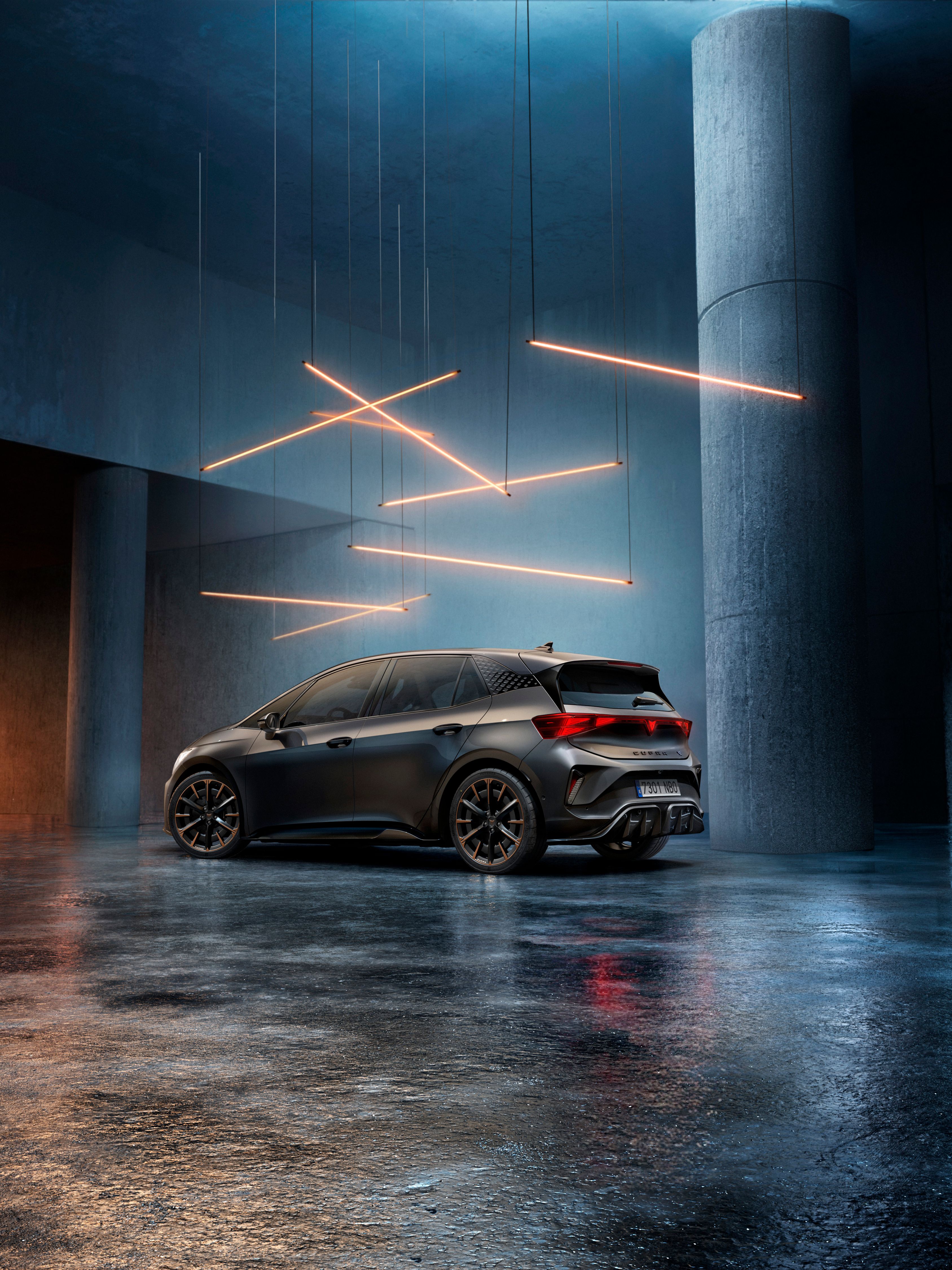 CUPRA Born stojí v štýlovo osvetlenom priemyselnom priestore s betónovými stenami a stĺpmi. Zo stropu visí niekoľko lineárnych svetelných zdrojov, ktoré vytvárajú elegantnú atmosféru. Vozidlo je vidieť zo zadnej strany a odráža svetlo na lesklej podlahe.