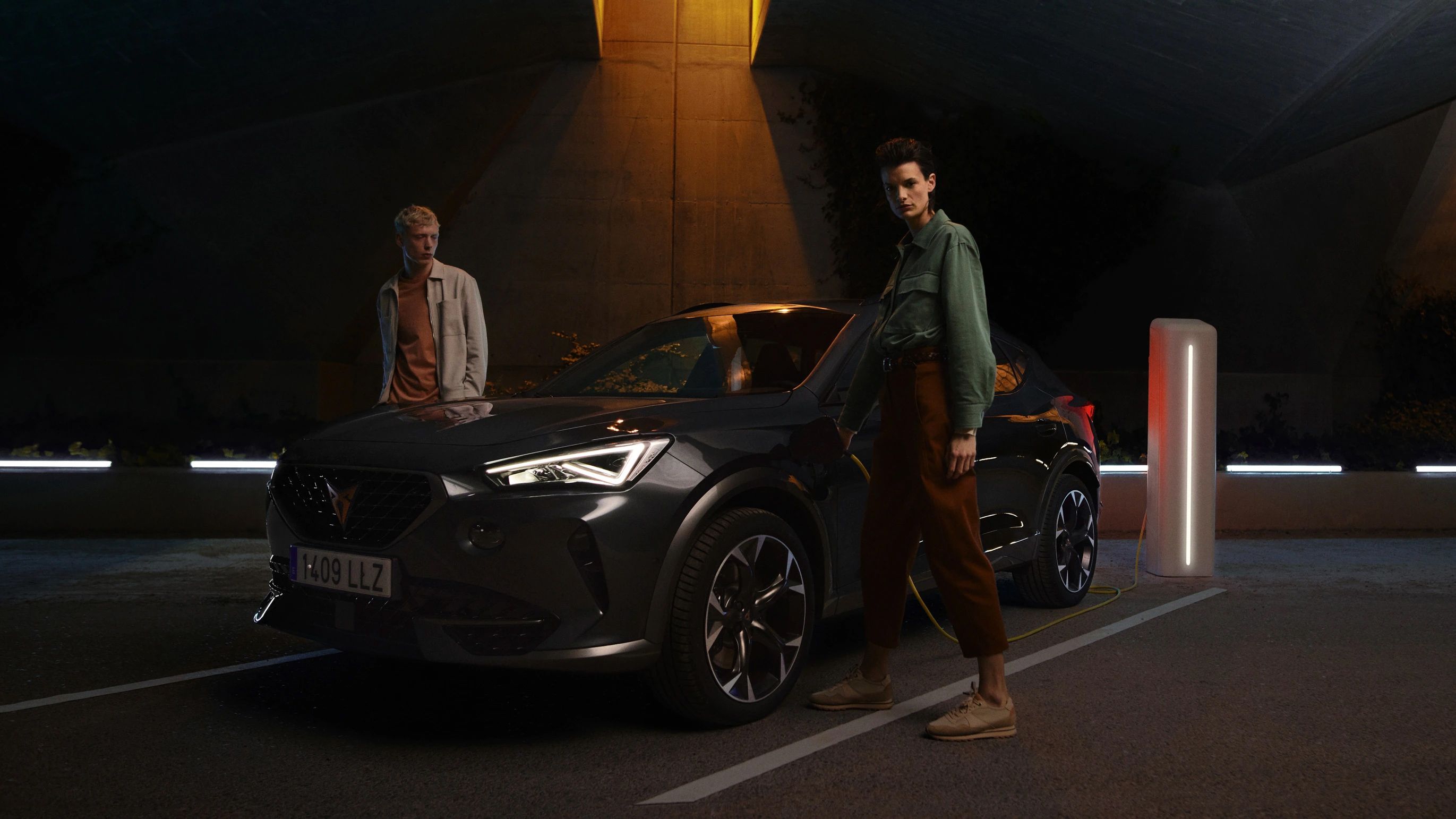 Dve osoby stoja pri tmavom aute, ktoré je zaparkované pod žiariacou lampou. Jedna osoba sa opiera o prednú časť auta, druhá stojí vedľa neho. Osvetlenie vytvára dramatickú atmosféru.