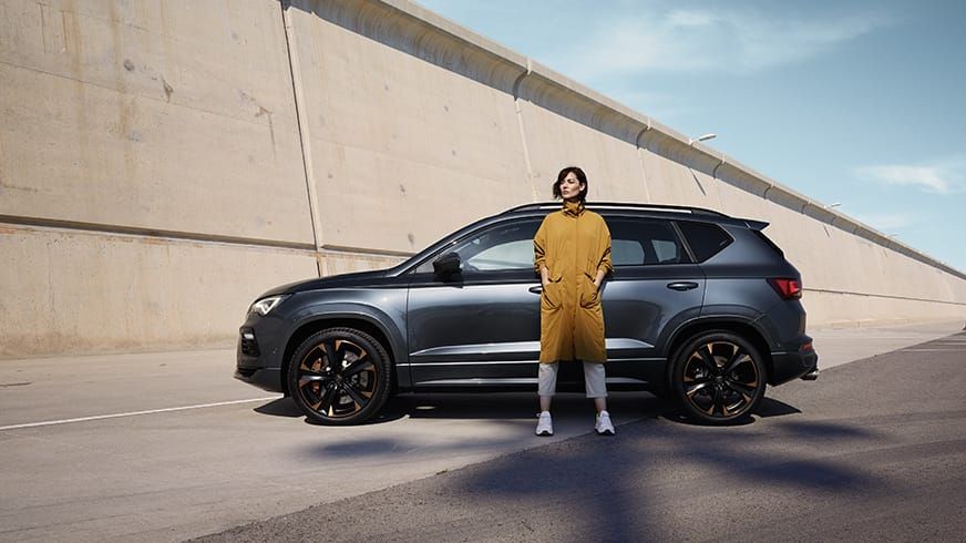 Žena v horčicovom kabáte stojí pred tmavosivým SUV na betónovej ceste. Pozadie tvoria betónové steny a zatiahnutá obloha.