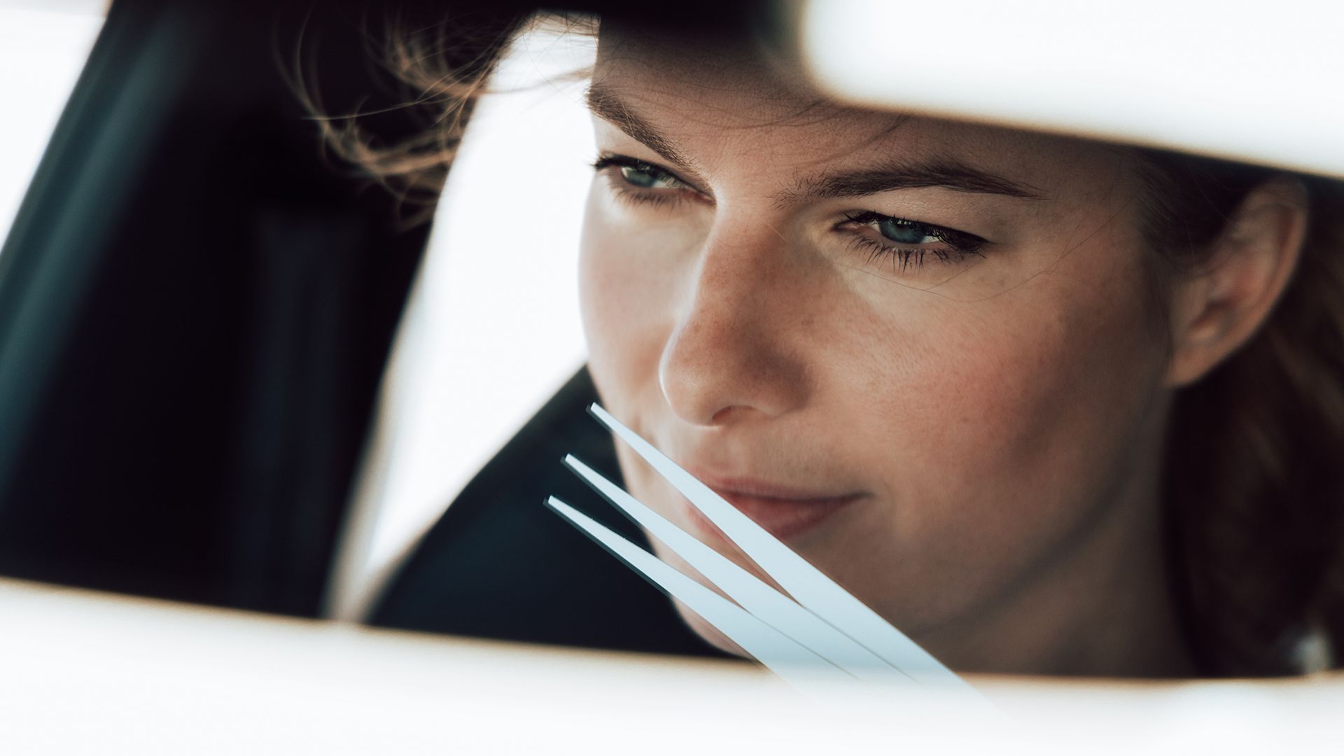 Žena sedí v aute. Má hnedé vlasy. Jej obraz zreteľne vidieť v spätnom zrkadle.