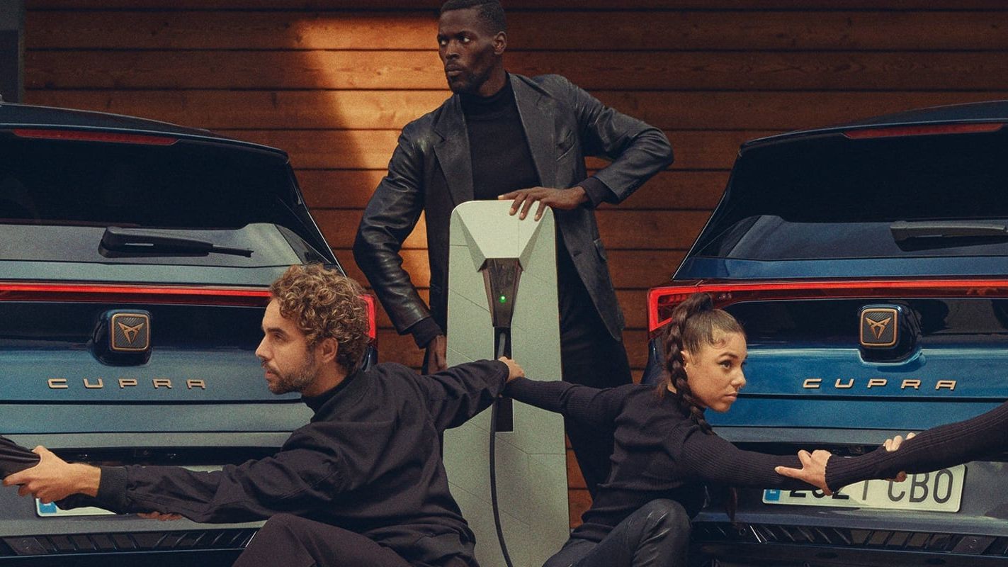 Skupina štyroch osôb v čiernom drží navzájom svoje ruky pred dvomi modrými autami Cupra. Postava v strede stojí, zvyšok sedí na zemi. 
