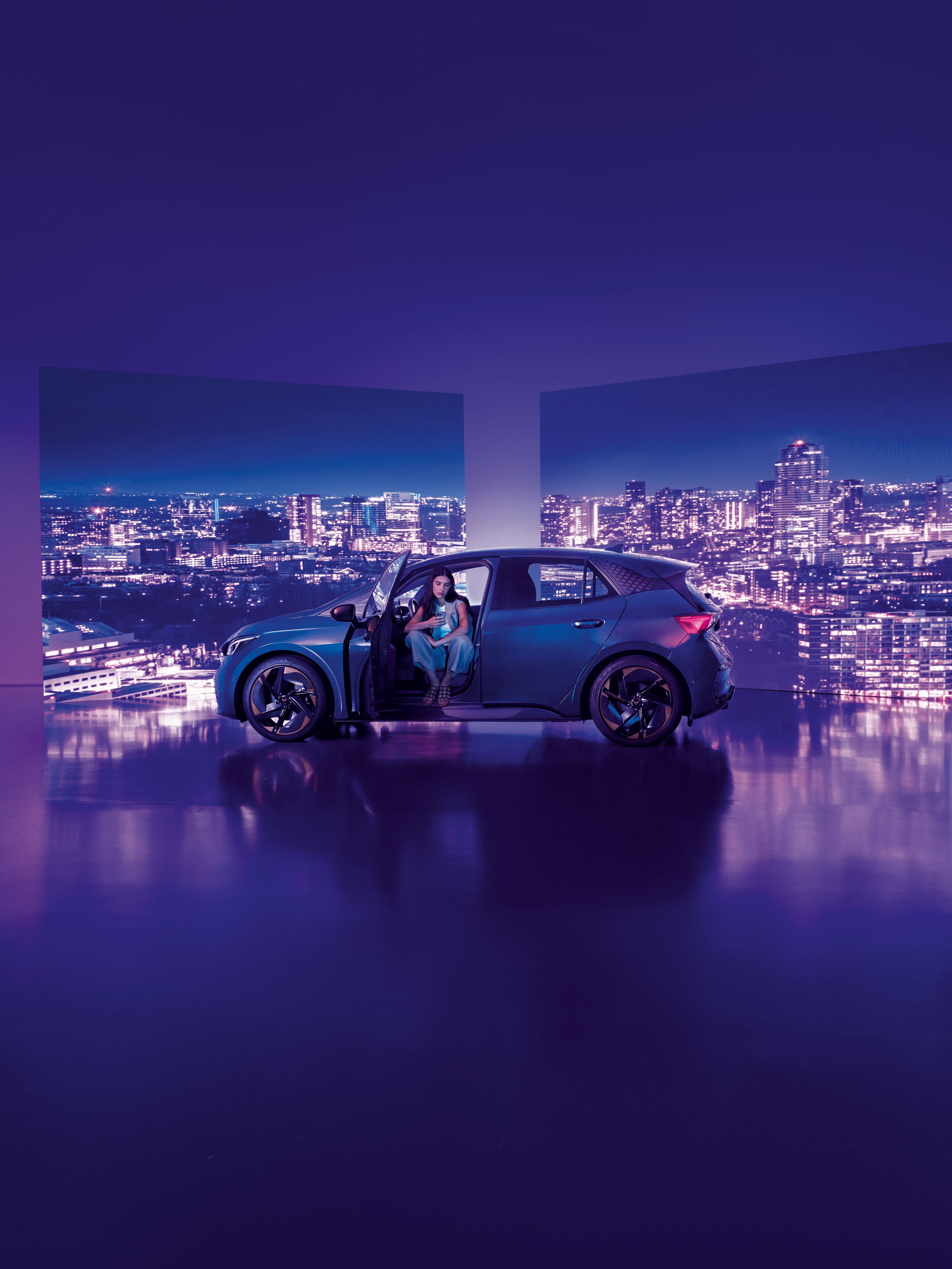 Pozadie tvoria dva veľké billboardy s panoramatickým výhľadom na nočné mesto, pred ktorými stojí moderné auto s otvorenými dverami. Žena sedí v aute na mieste vodiča, interiér je osvetlený jemným svetlom.