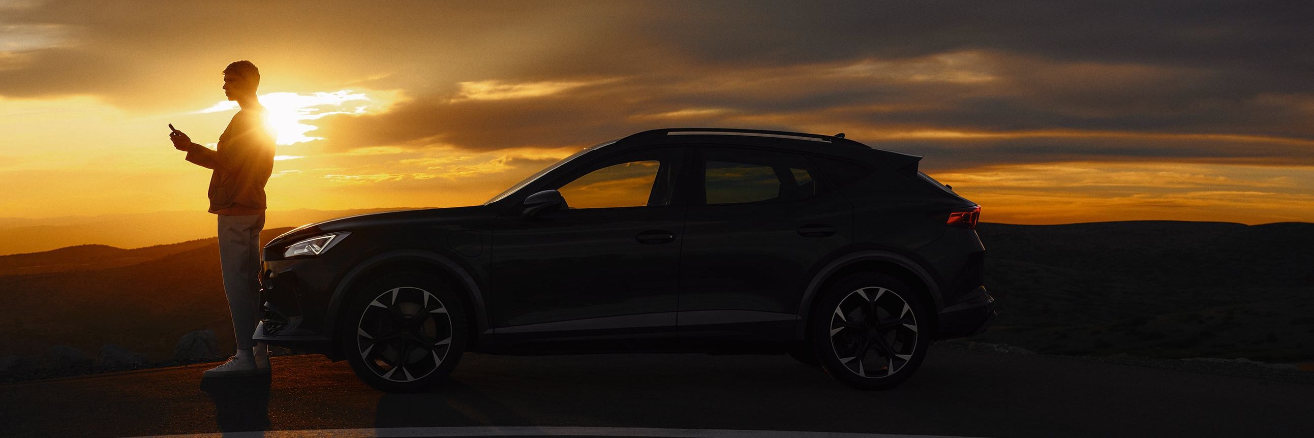 Osoba stojí vedľa zaparkovaného automobilu na kraji cesty pri západe slnka. Obloha je pokrytá tmavými mrakmi, ktoré vytvárajú dramatický efekt svetla. 