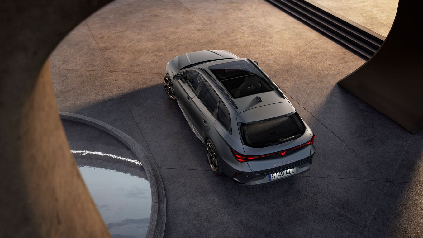 Šedé športové auto stojace na betónovej ploche pri modernej stavbe. Auto je zobrazené z vtáčej perspektívy. Vedľa neho vidieť kruh, v ktorom je voda.