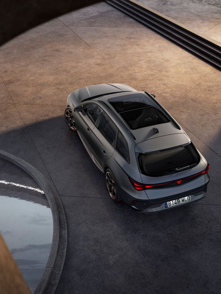 Šedé športové auto stojace na betónovej ploche pri modernej stavbe. Auto je zobrazené z vtáčej perspektívy. Vedľa neho vidieť kruh, v ktorom je voda.