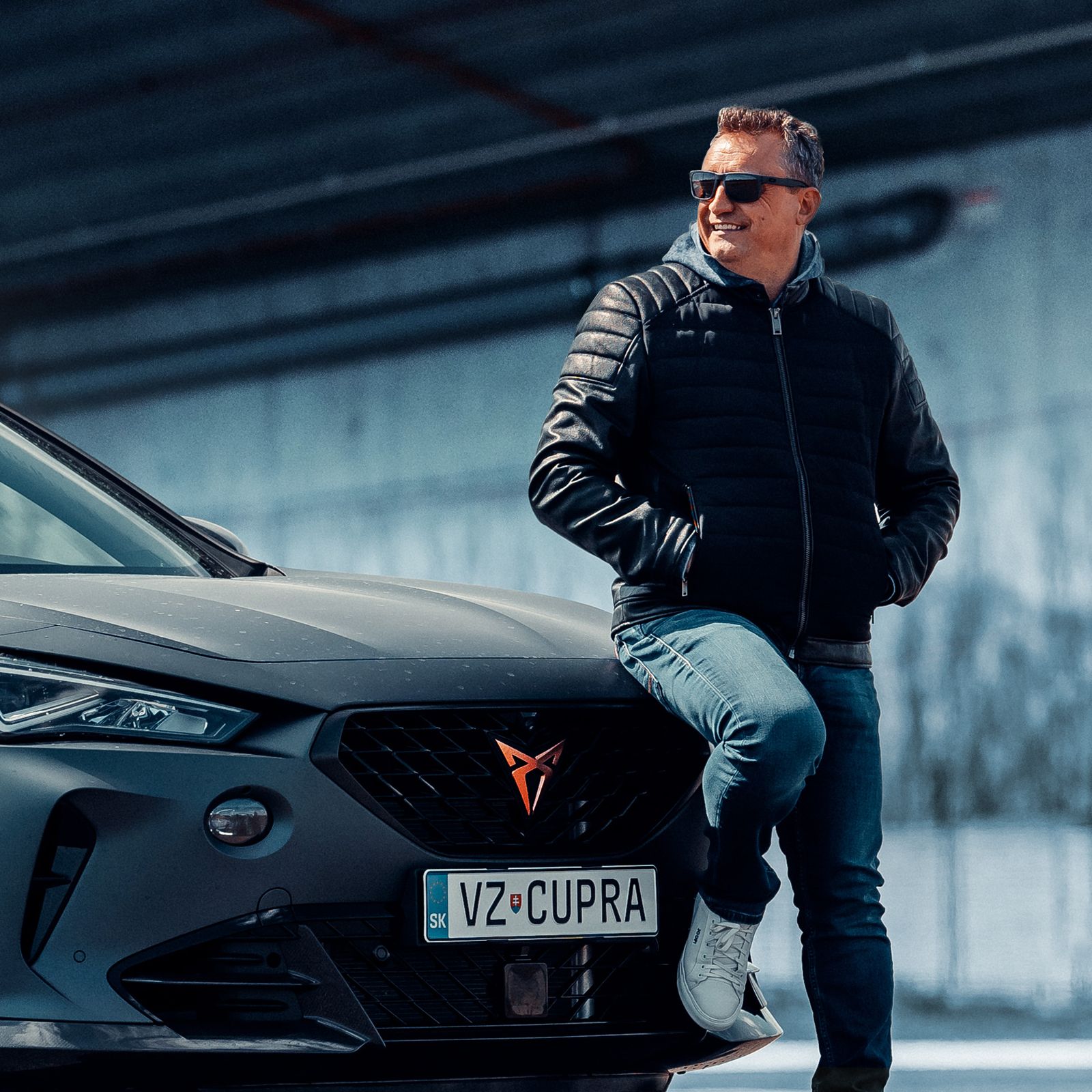 Muž opretý o prednú časť sivého športového auta značky CUPRA s evidenčným číslom VZ CUPRA, na pozadí moderná architektúra. Muž má na sebe tmavú bundu a modré džínsy.