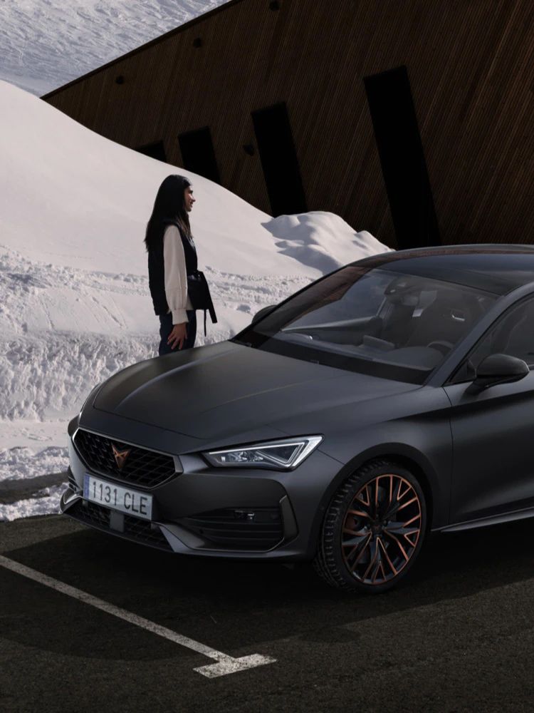 Na obrázku je zaparkované tmavo sivé auto, ktoré stojí pred drevenou budovou pri zasneženom svahu. Vedľa auta stojí osoba. 