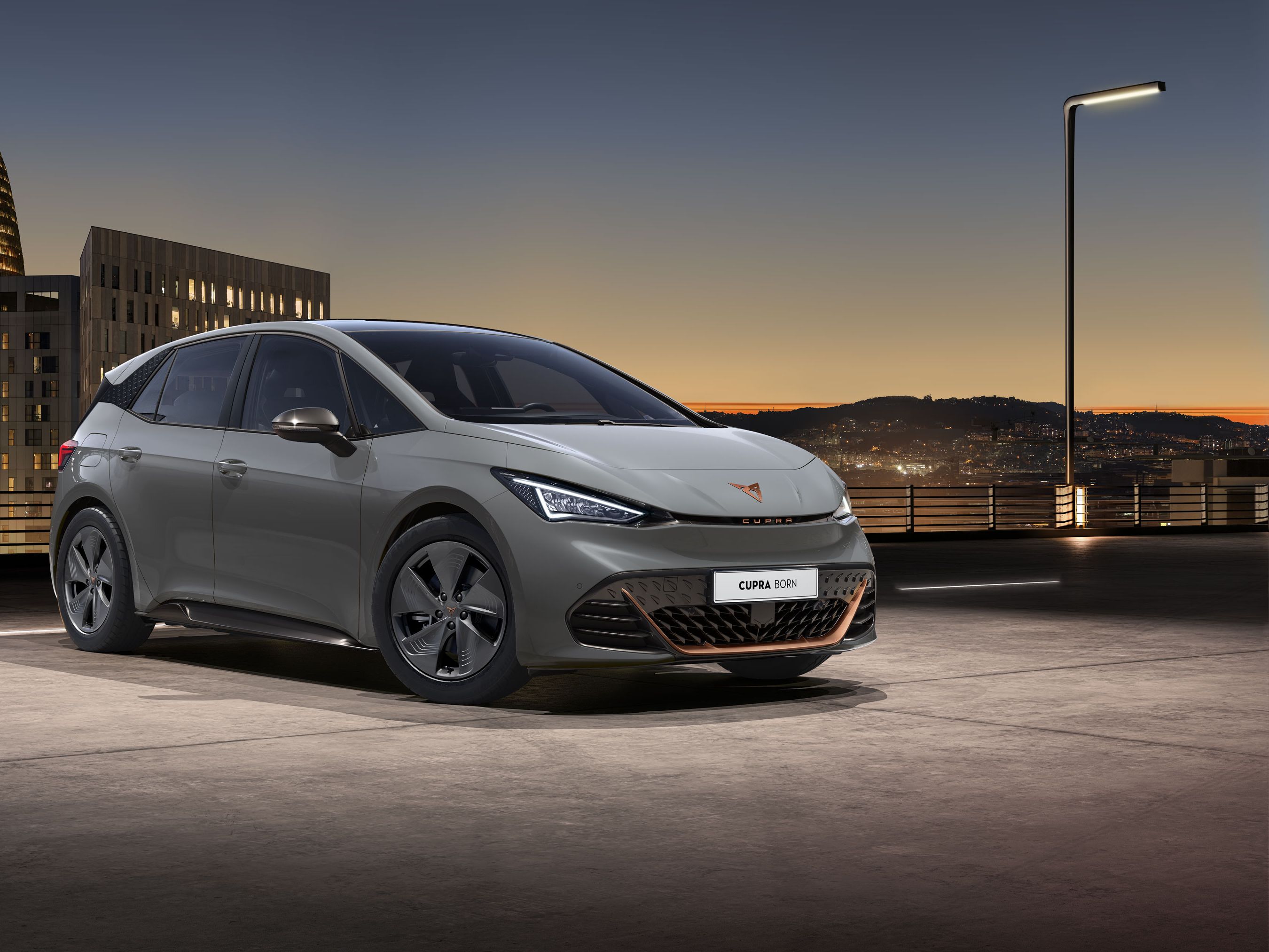 CUPRA Born v sivom lakovaní zaparkované na streche parkovacieho domu s výhľadom na výškové budovy centra mesta.