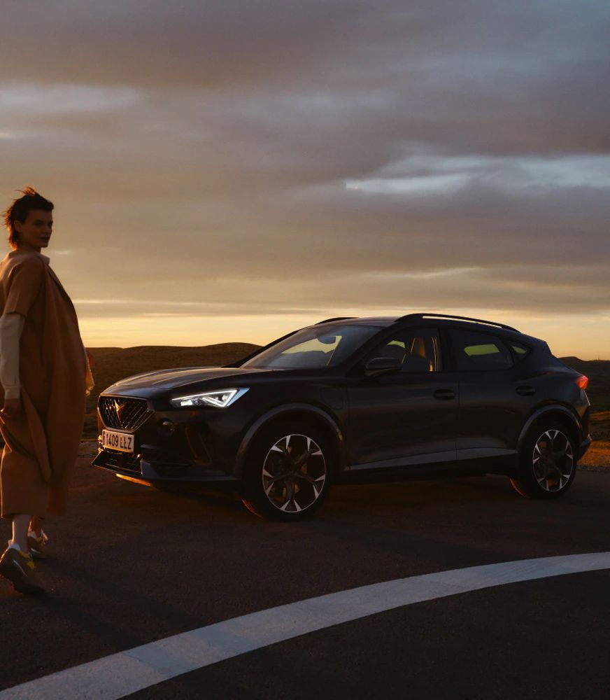 Žena stojí vedľa zaparkovaného športového auta na ceste počas západu slnka. Krajina v pozadí je osvetlená teplými farbami.